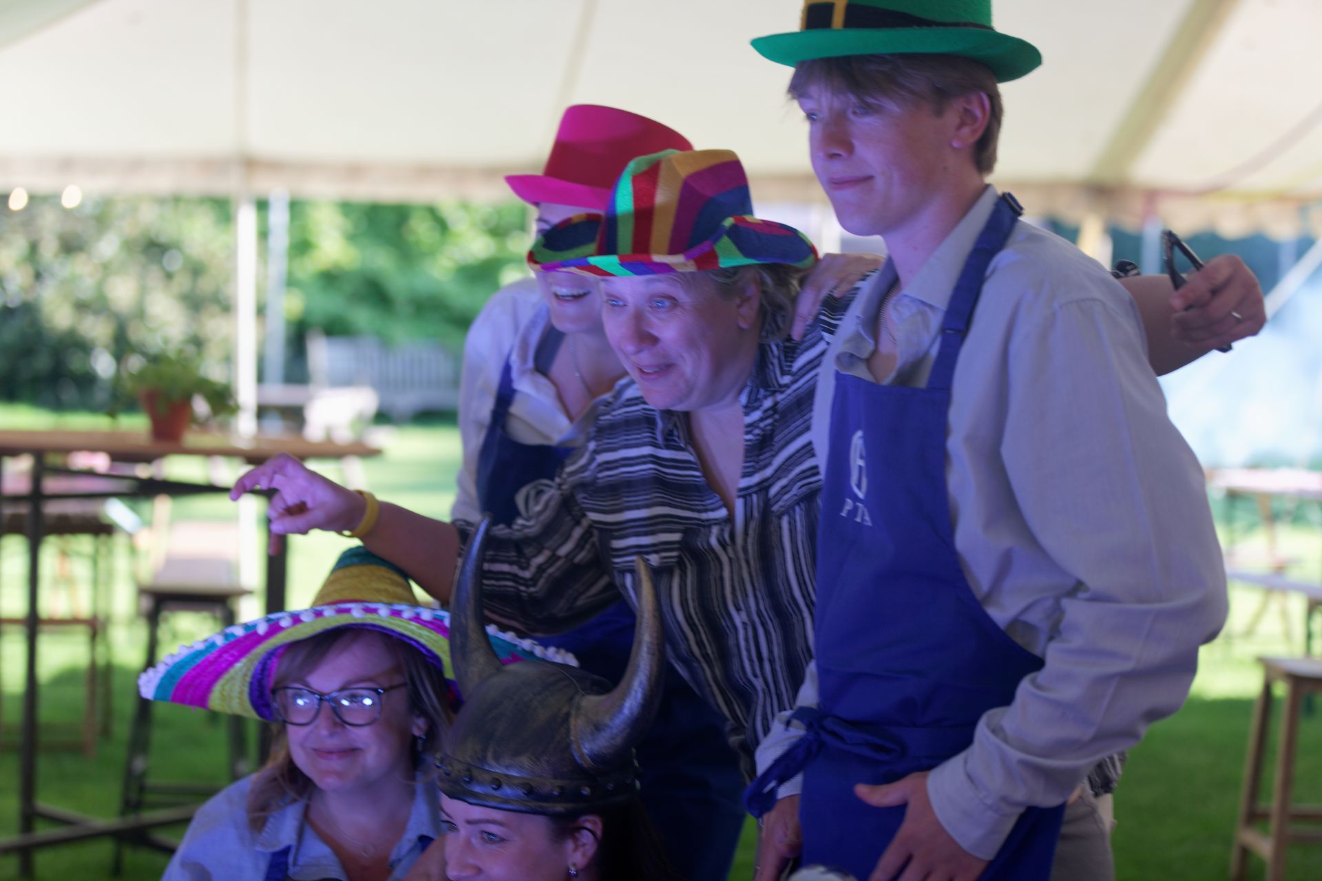 Group of people in hats and aprons pose in front of a tent. Some point and smile.