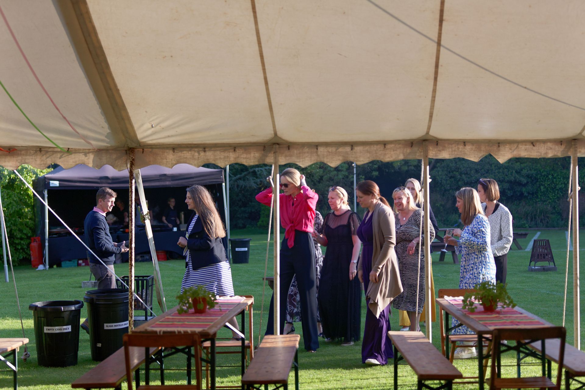 People in a tented outdoor event. Tables, benches, and a catering setup are visible.