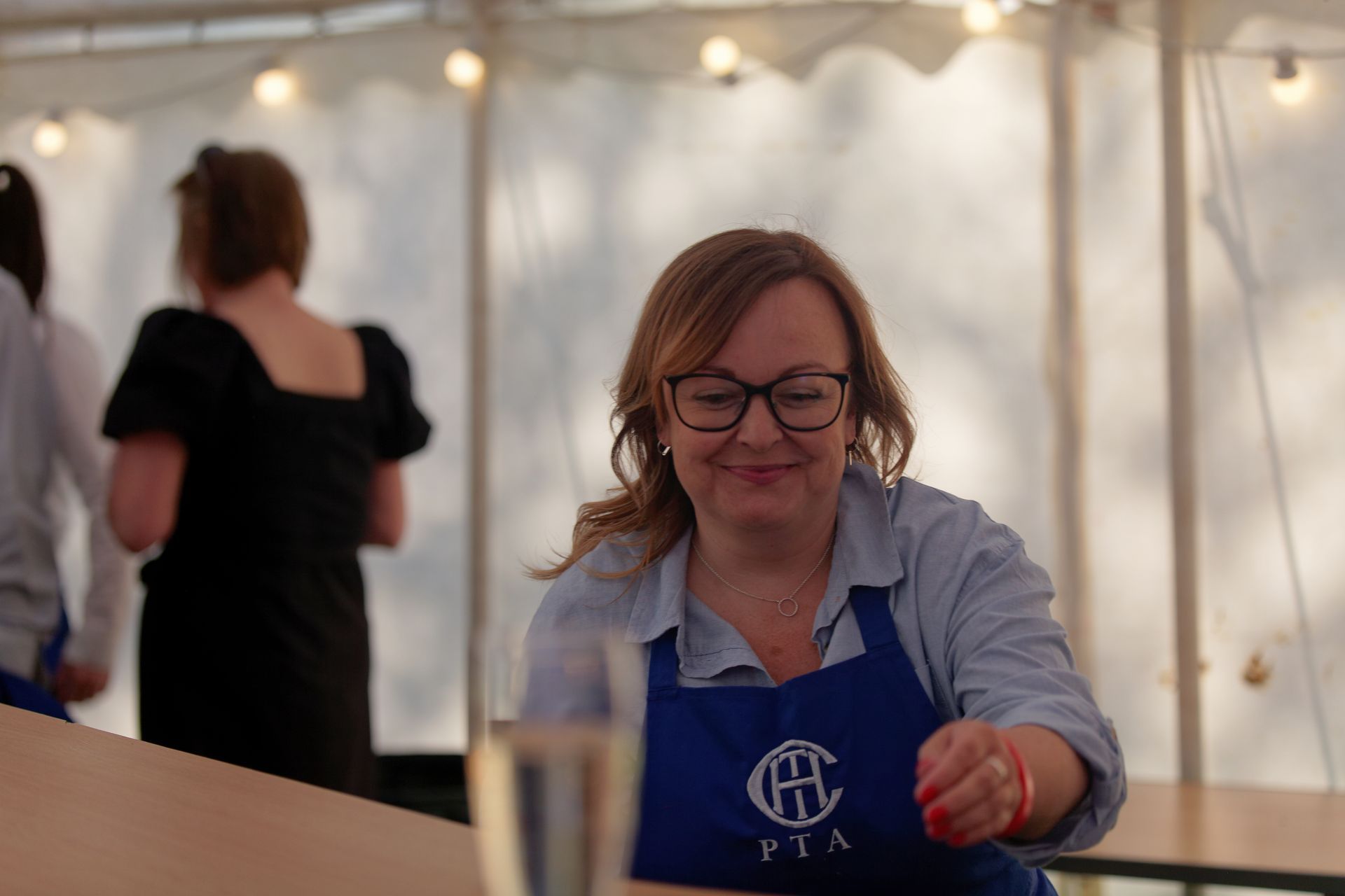 Woman in glasses and apron smiles, possibly at a PTA event. Inside tent, other people are blurred in the background.