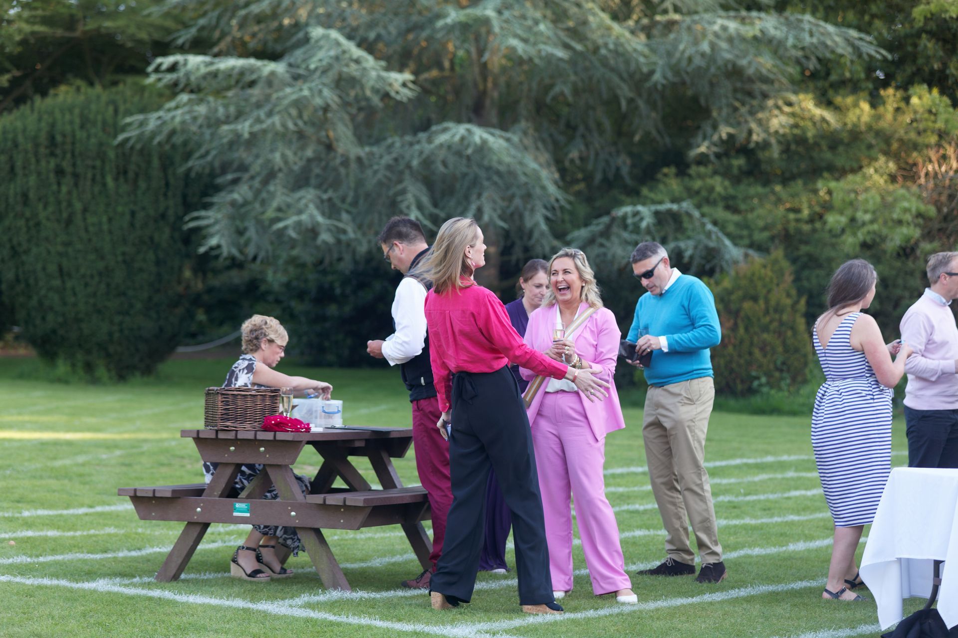 People socializing outdoors on a lawn. A woman in pink pantsuit laughs. Others are near a picnic table.
