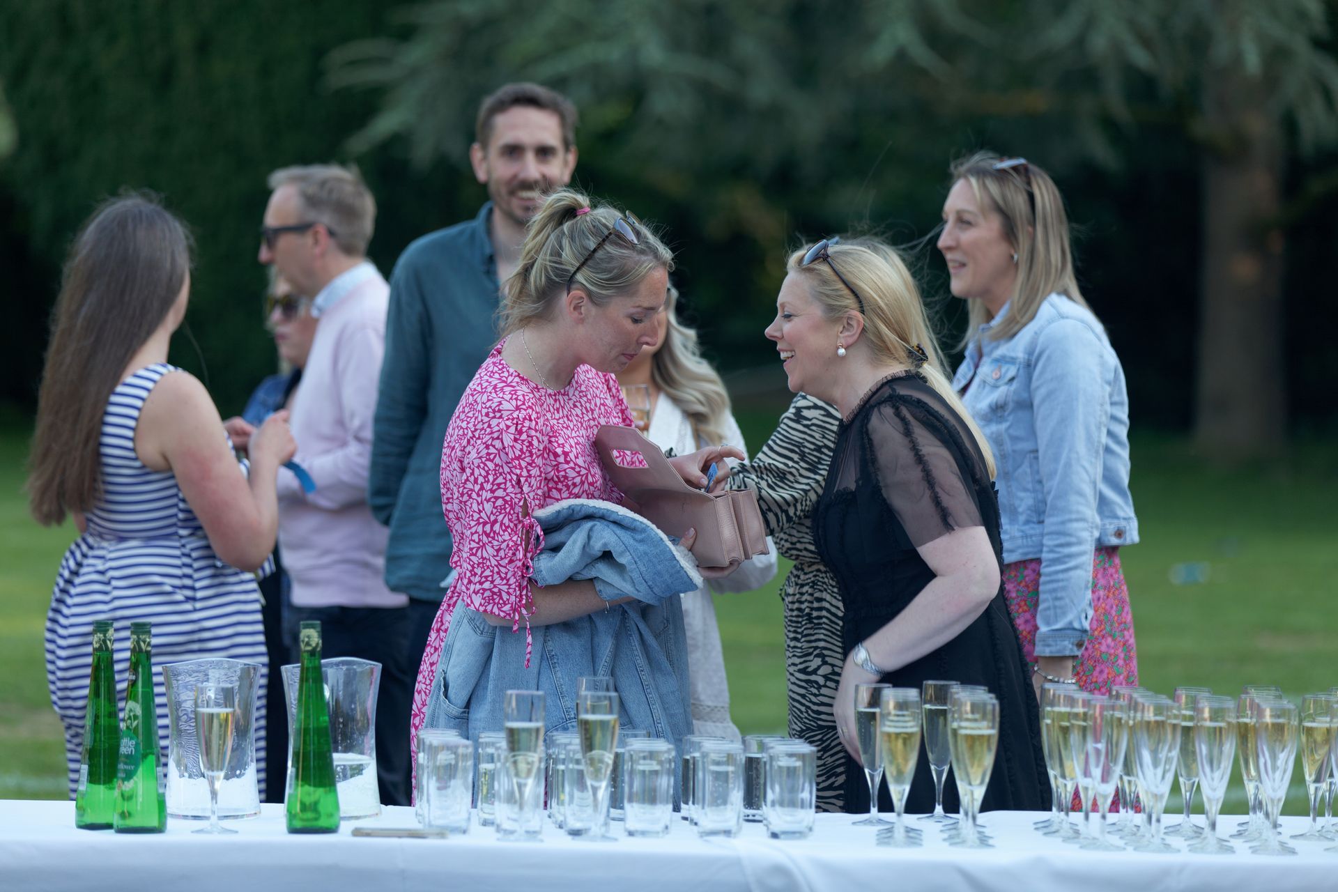 Group of people at an outdoor gathering, laughing and talking near a table of drinks.