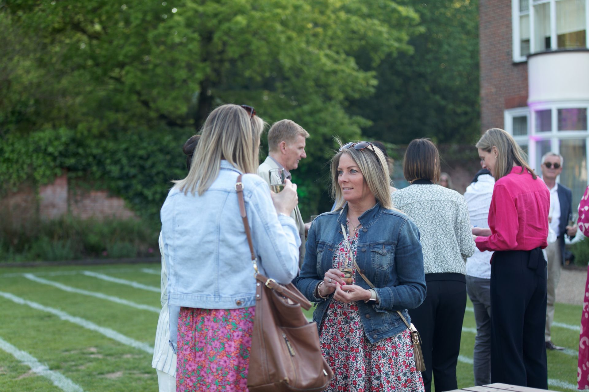 People socializing outdoors, woman in denim jacket. Grass lawn, building in background.