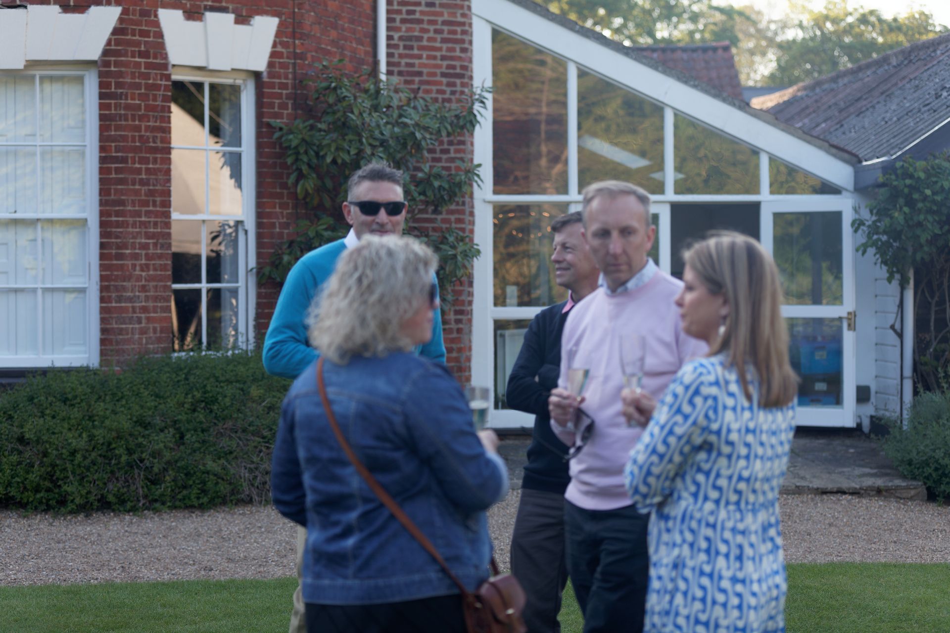 Group of people talking outside a house with large windows, green grass, and trees.