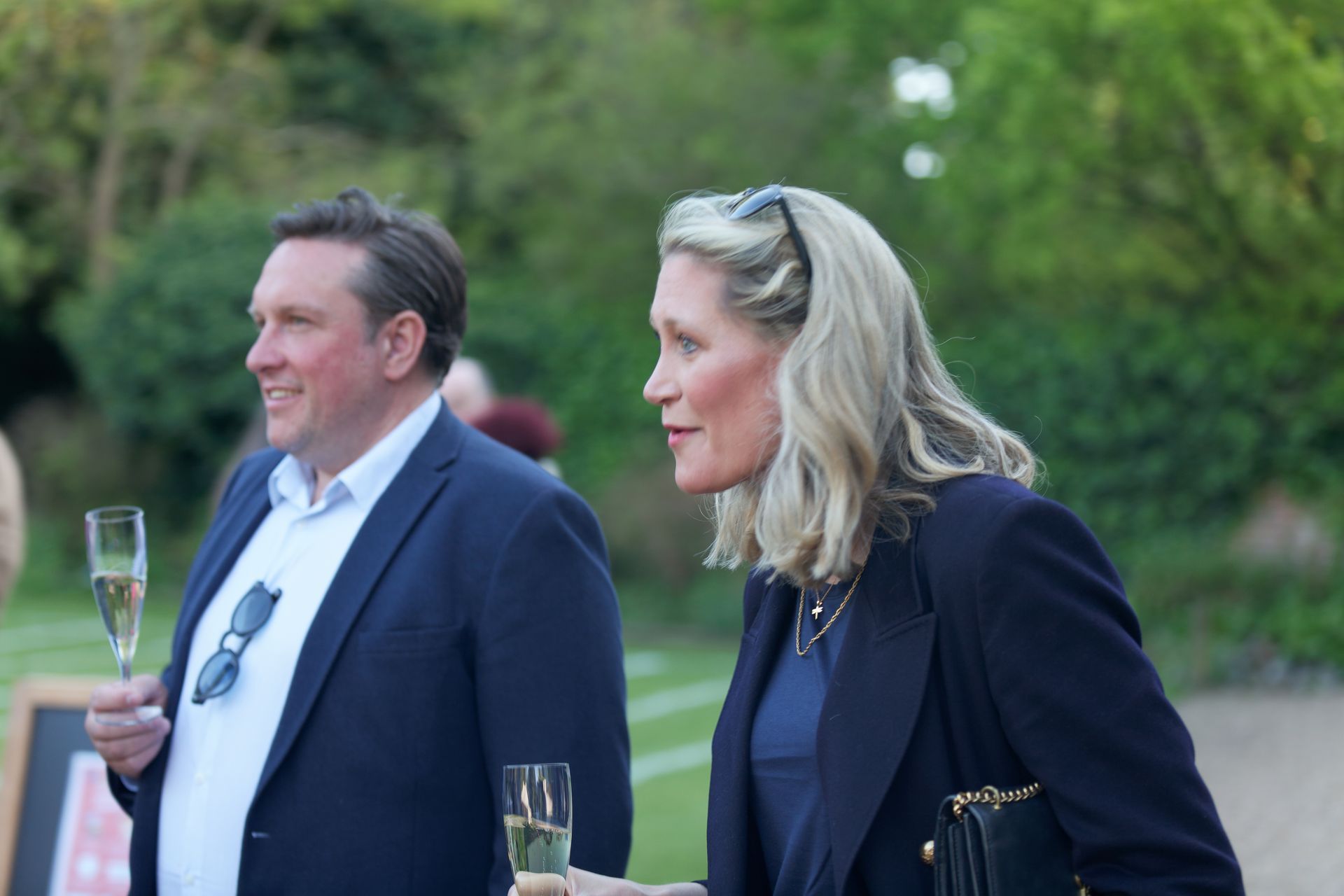 Man and woman in navy blazers, holding champagne flutes, smiling outdoors at an event.