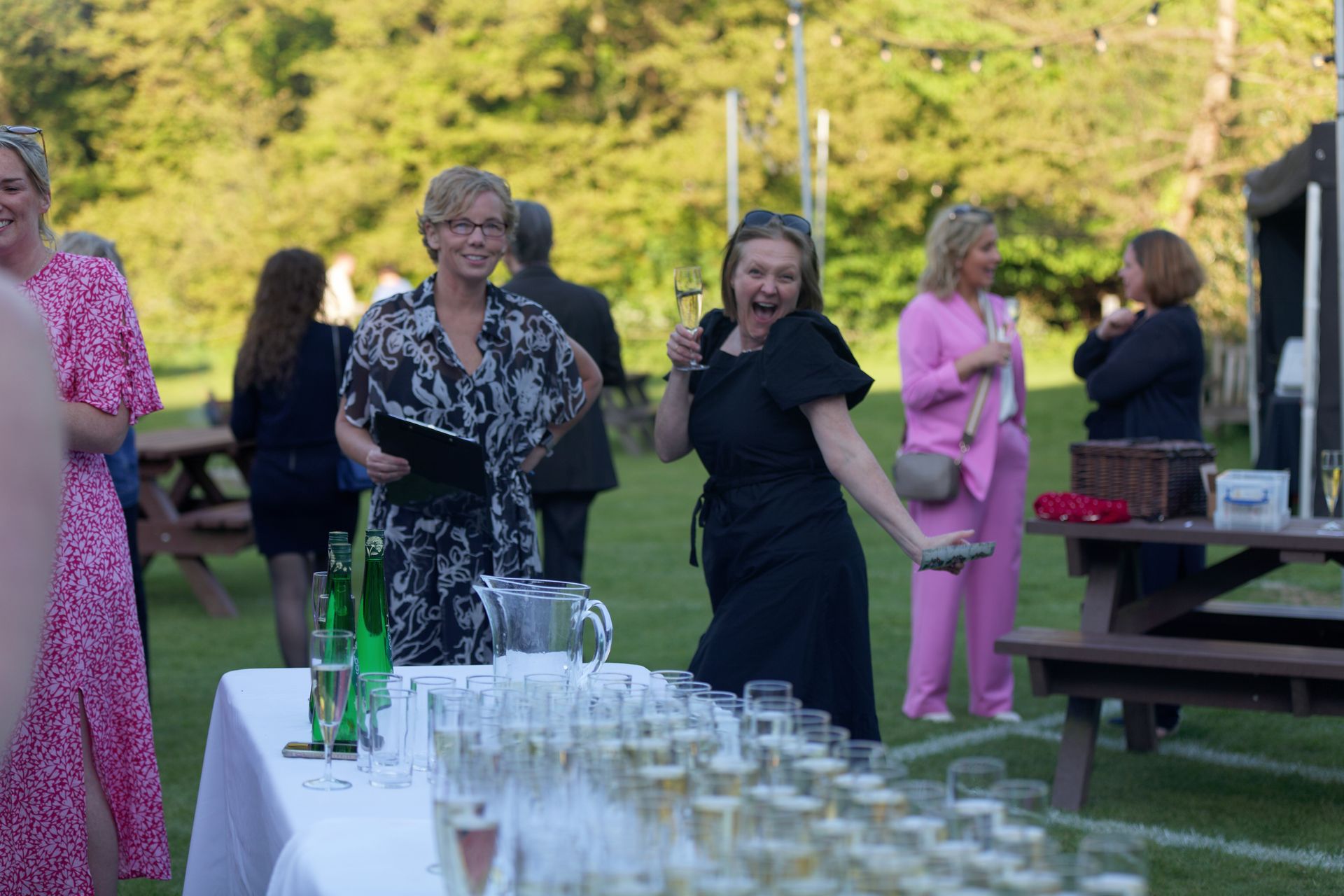 People at an outdoor event, some holding drinks. A woman in a black dress is excited, near a table of champagne glasses.
