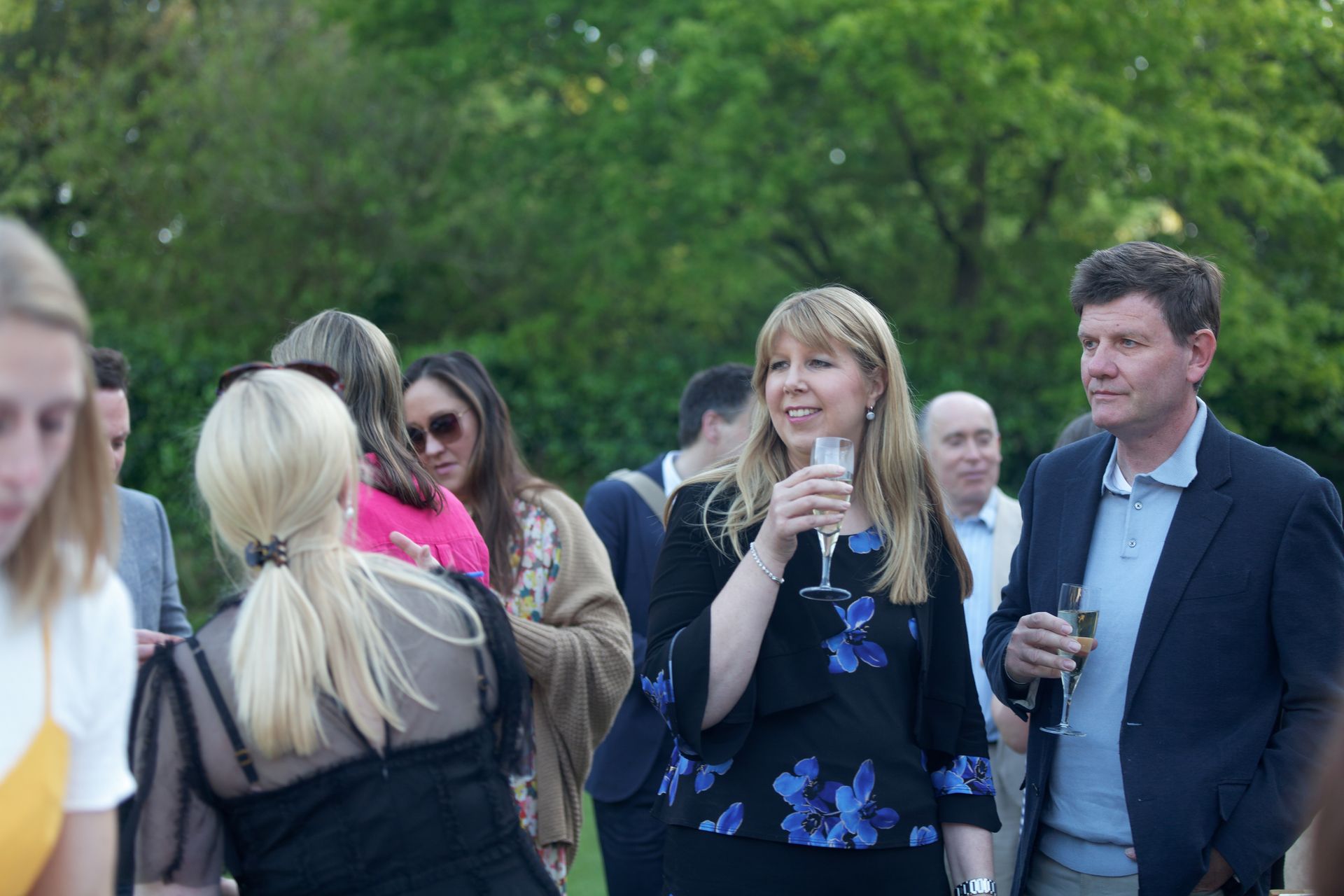 People at an outdoor event; woman sips champagne, man holds a drink. Green grass and trees in background.