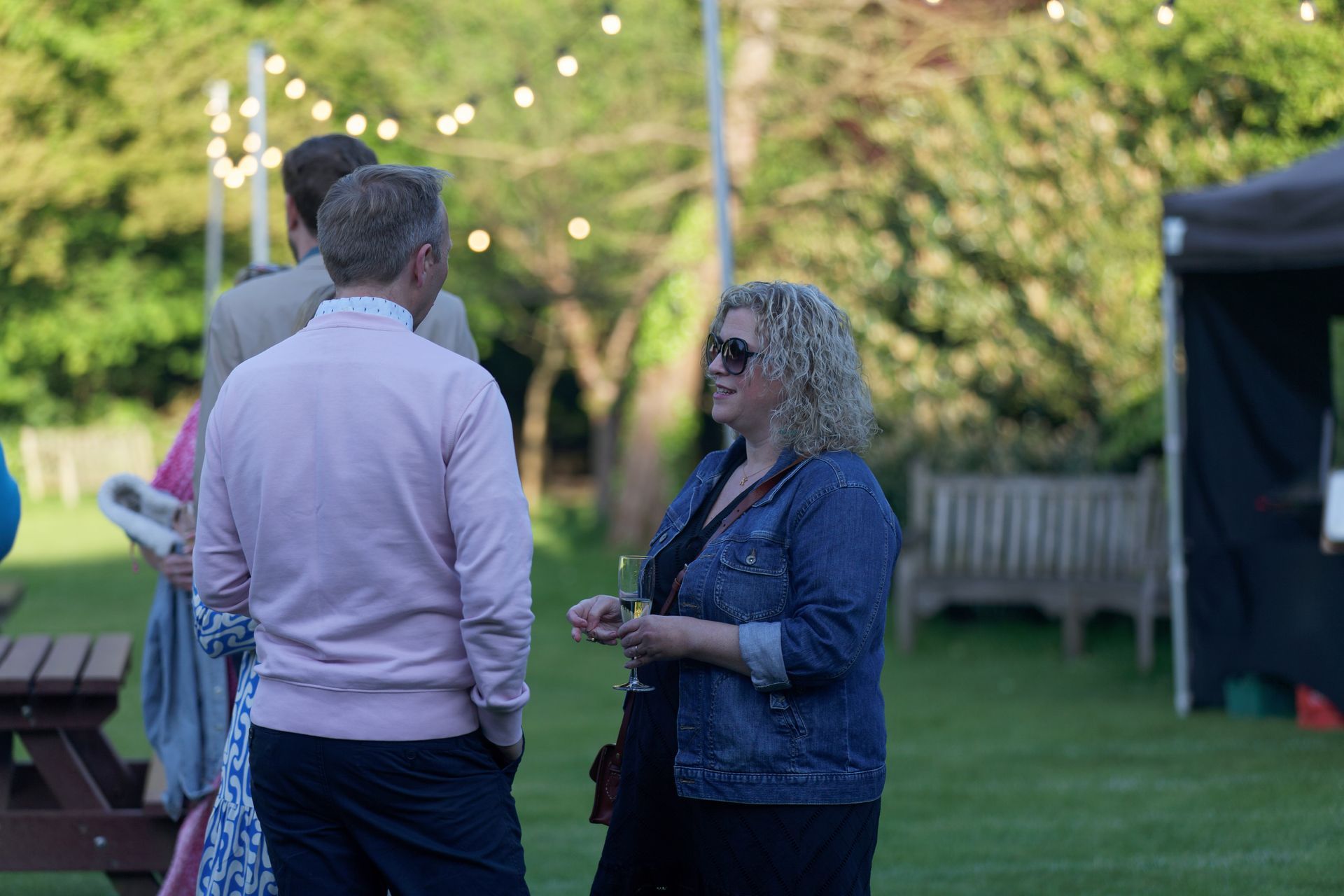 People in a garden, socializing. Woman in a denim jacket, talking to a man in a pink sweater. Party lights overhead.