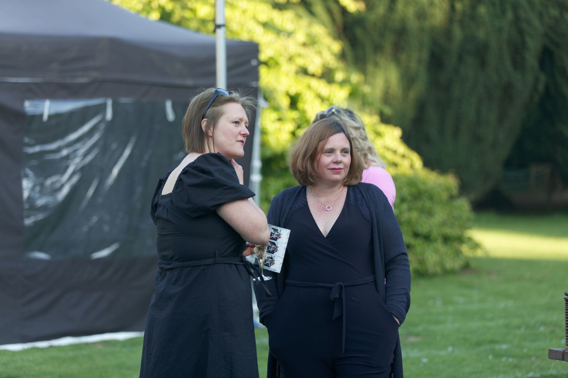 Two women in black dresses stand on a lawn near a black tent; one holds a small item, chatting.