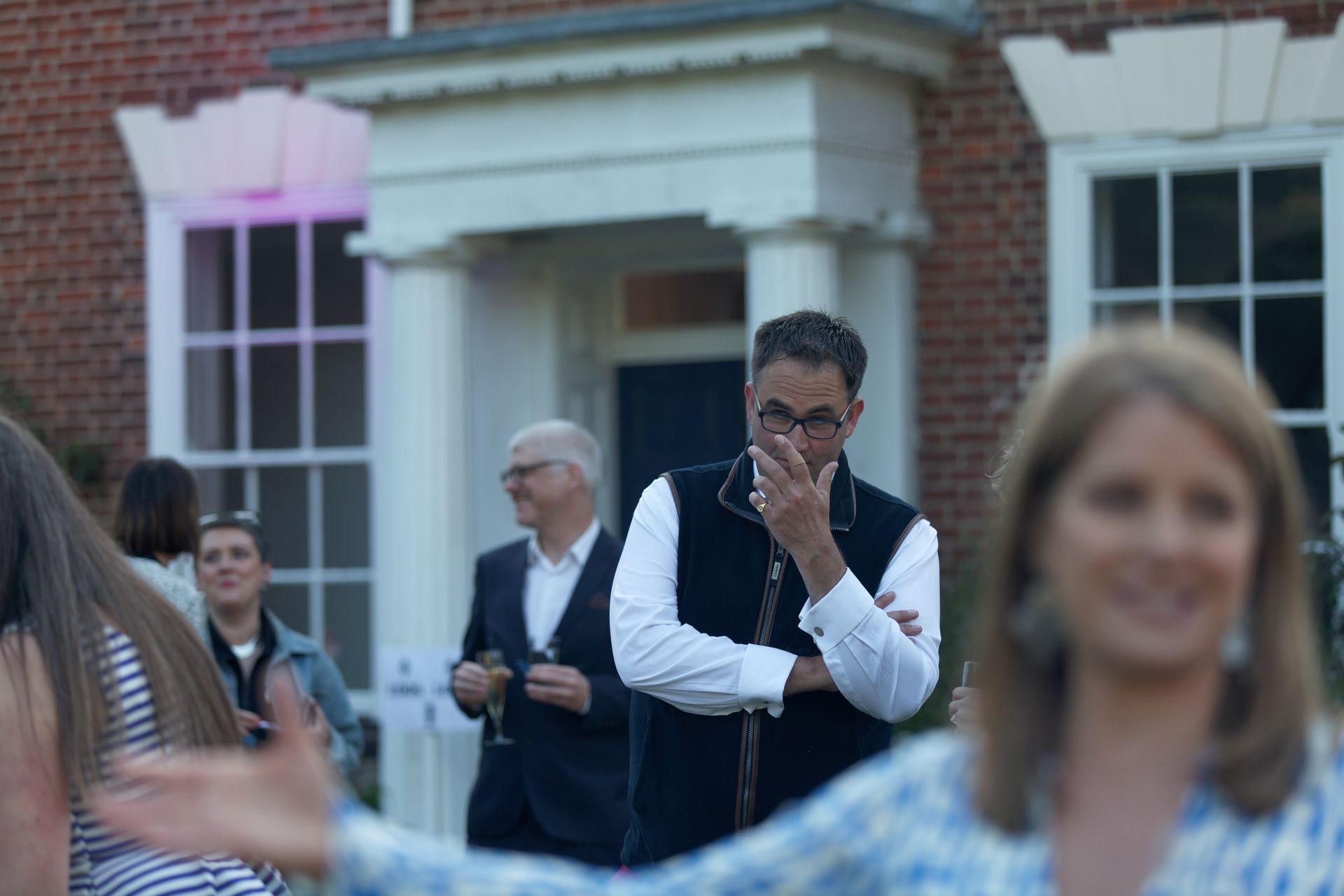 Man in glasses at outdoor event, touching face, with house in background and people.