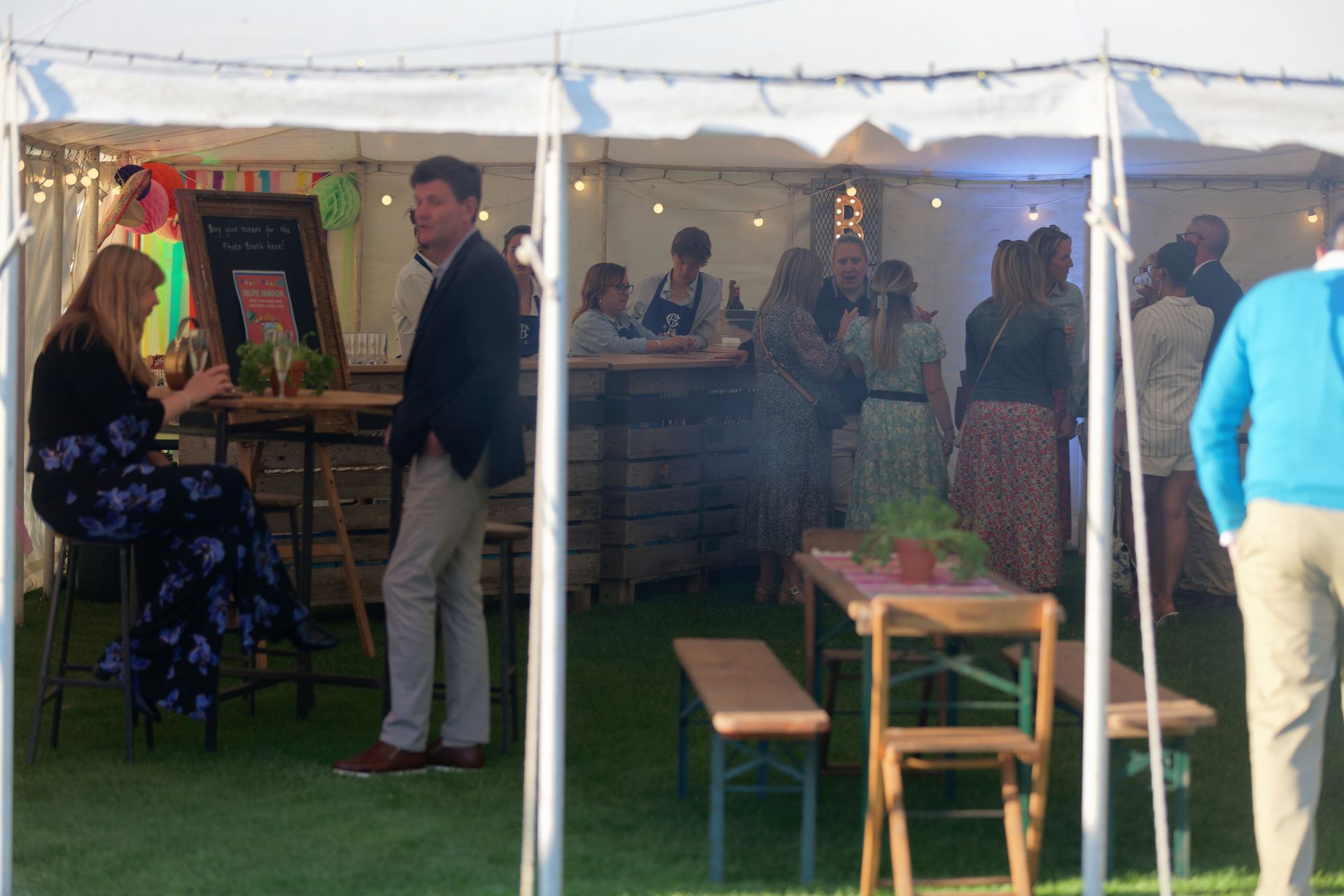 People socializing inside a white tent, including a man in a blazer and a woman in a floral dress, with a bar area.