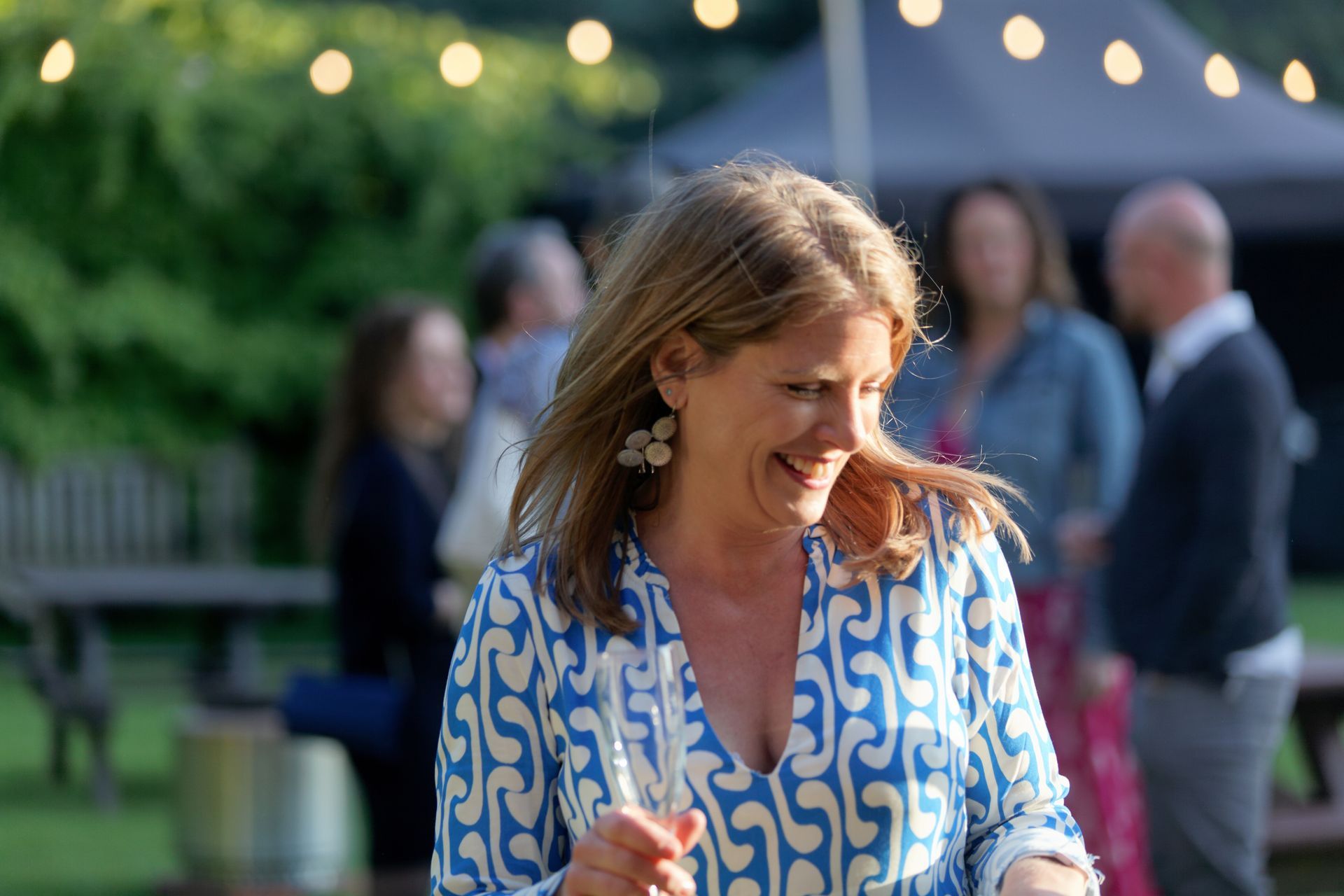 Woman in blue patterned dress laughs at an outdoor party, string lights overhead.