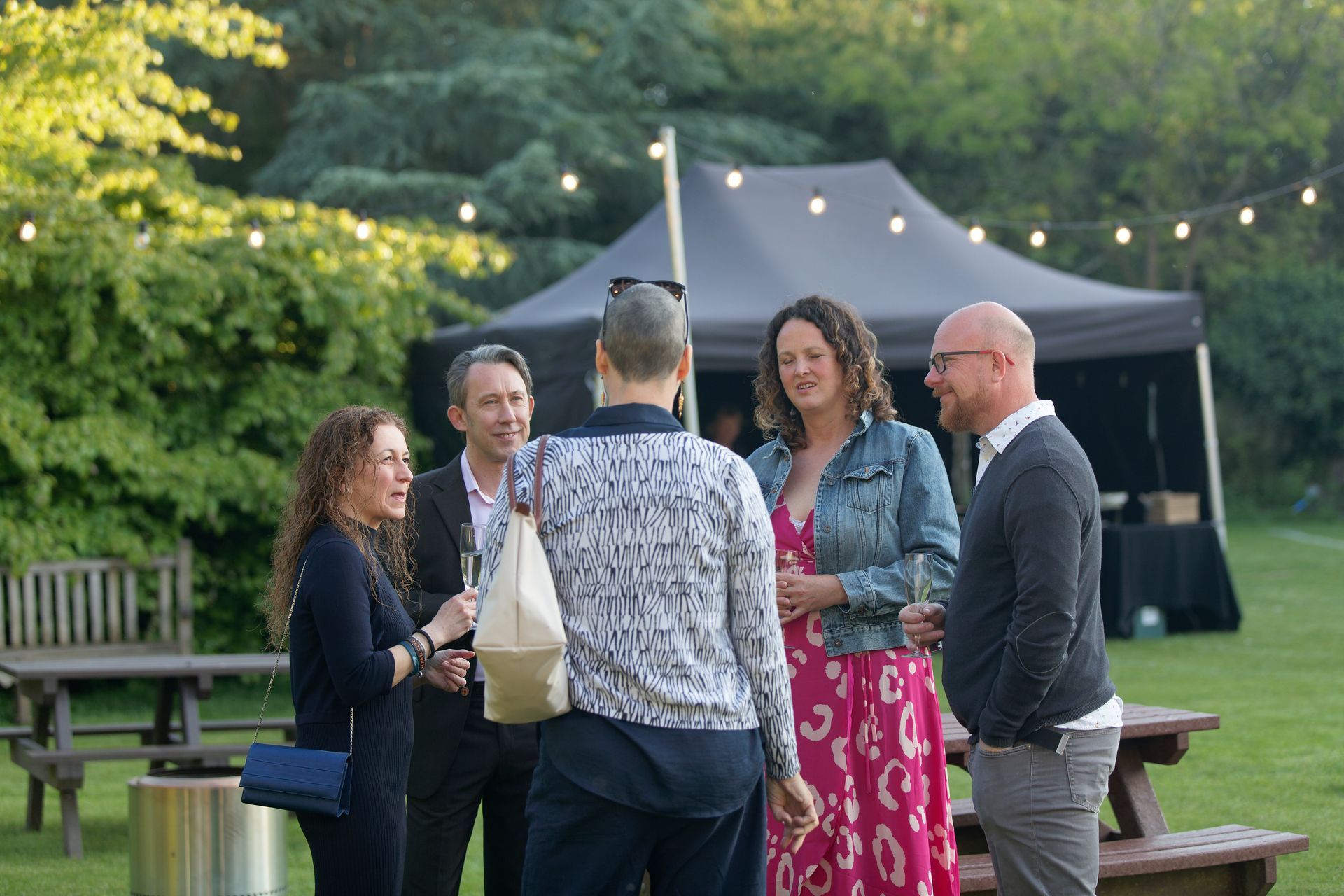 Group of six people chatting outdoors in a garden; a tent, string lights, and tables are visible.