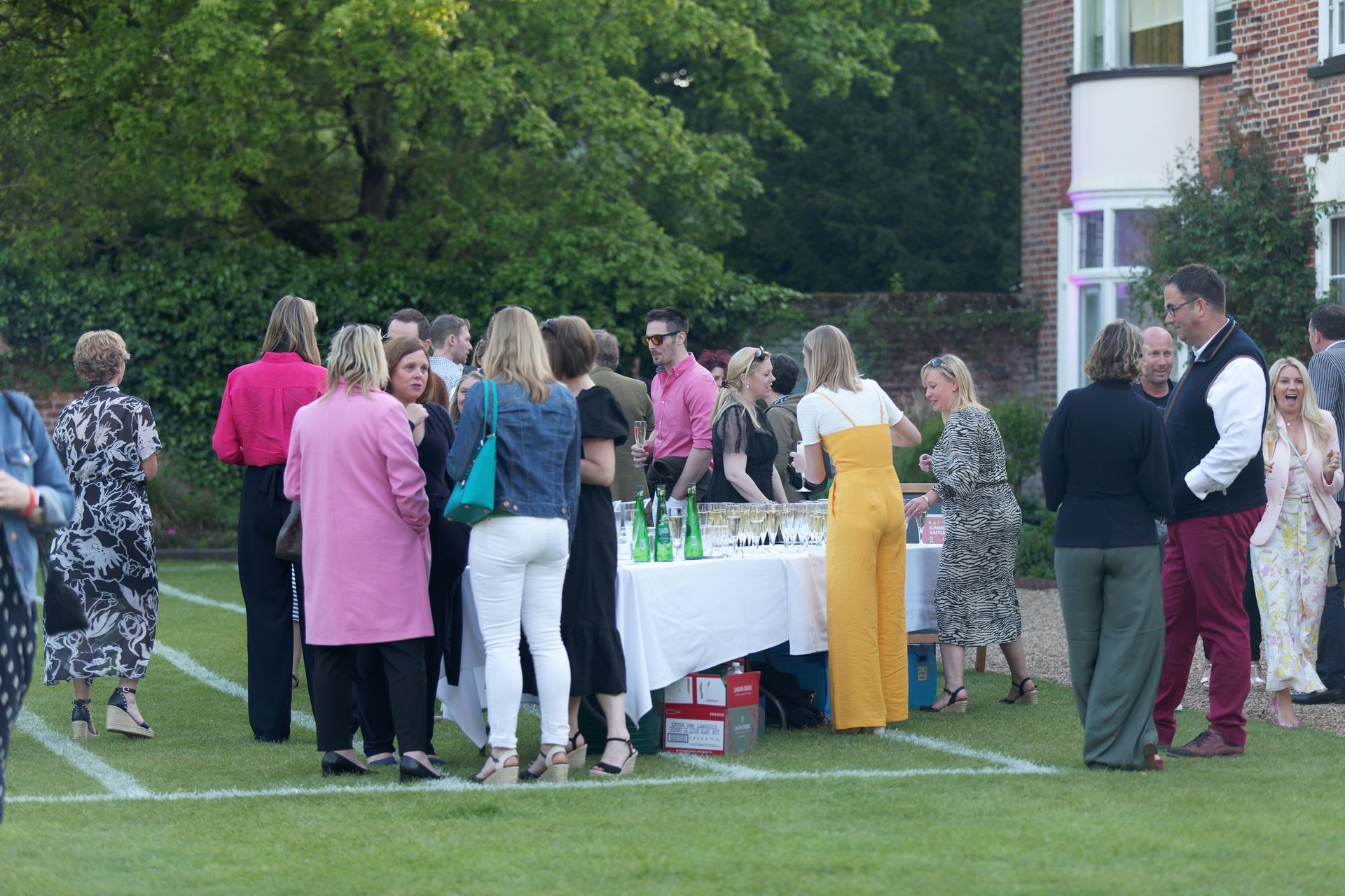 People gathered around a table on a lawn.  Some wearing pink, a building in the background.