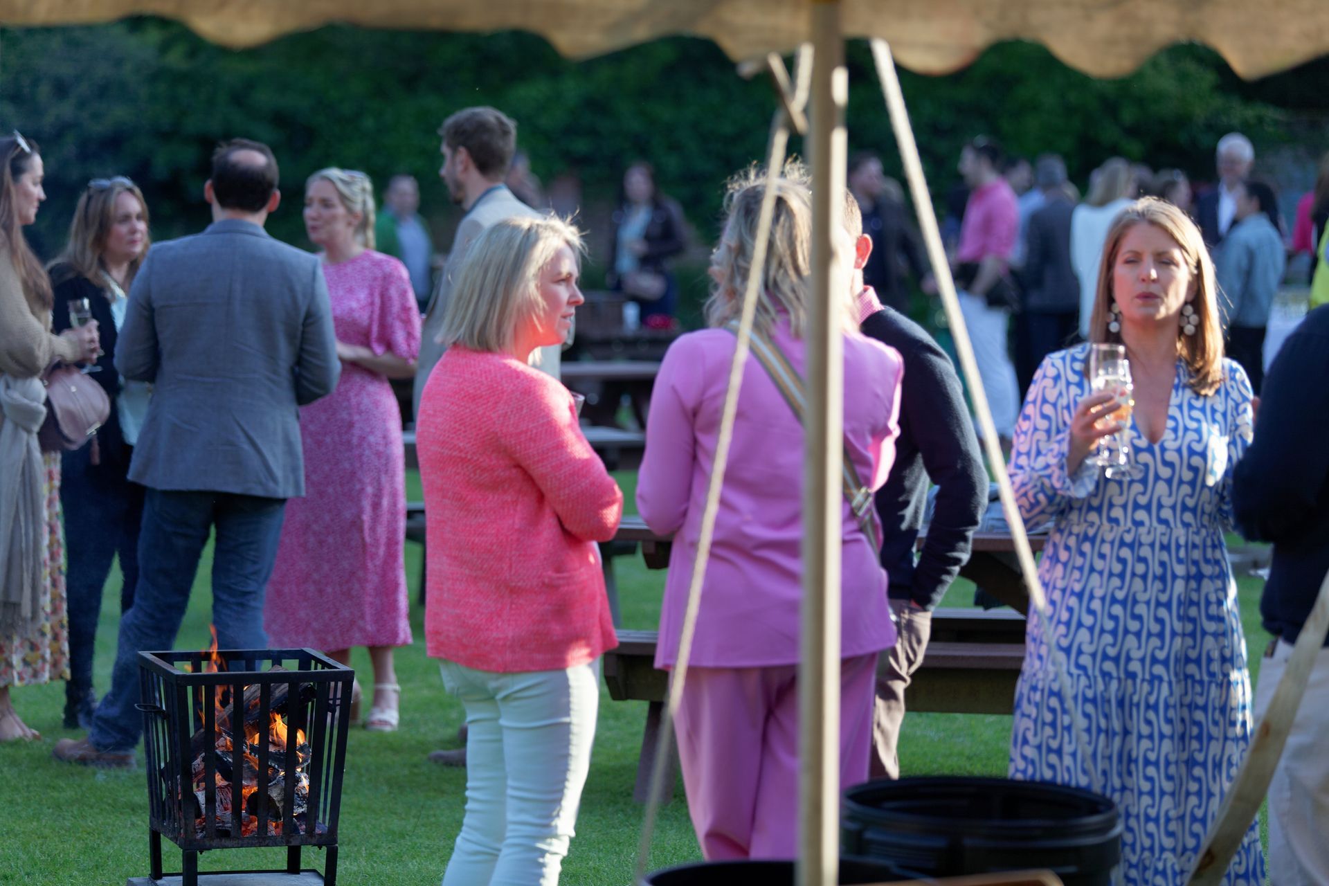 People socializing outdoors at an event, some holding drinks. Pink and blue outfits, fire pit, lawn setting.