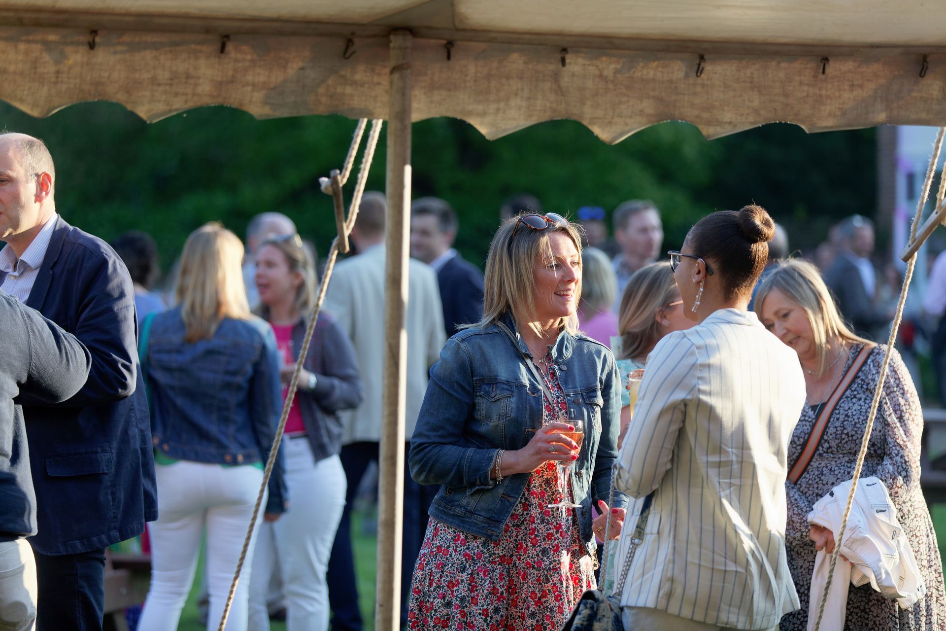 People socializing under a tent, some holding drinks. Green lawn, sunny day.