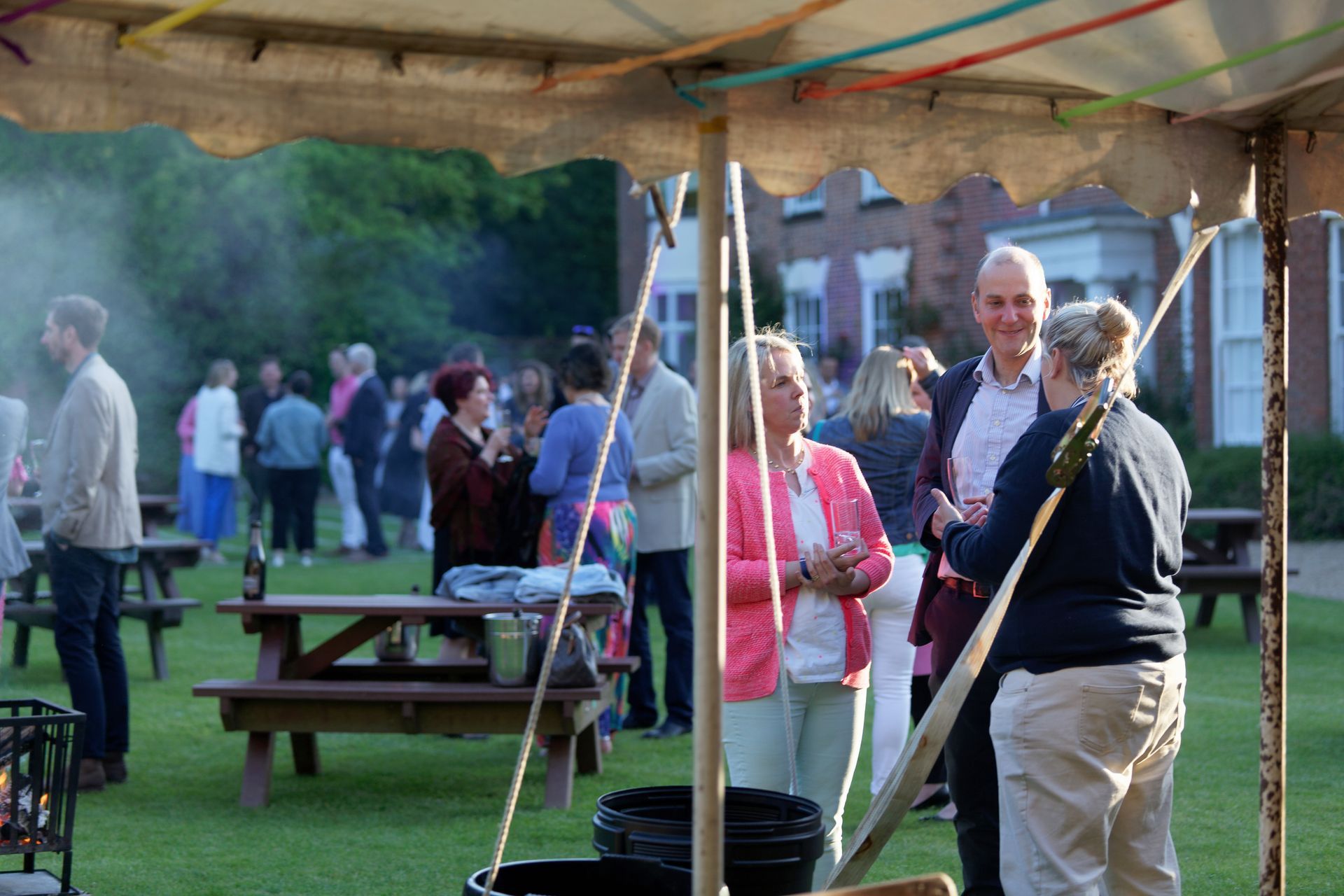 People socializing outdoors under a tent, chatting and mingling on a lawn with picnic tables.