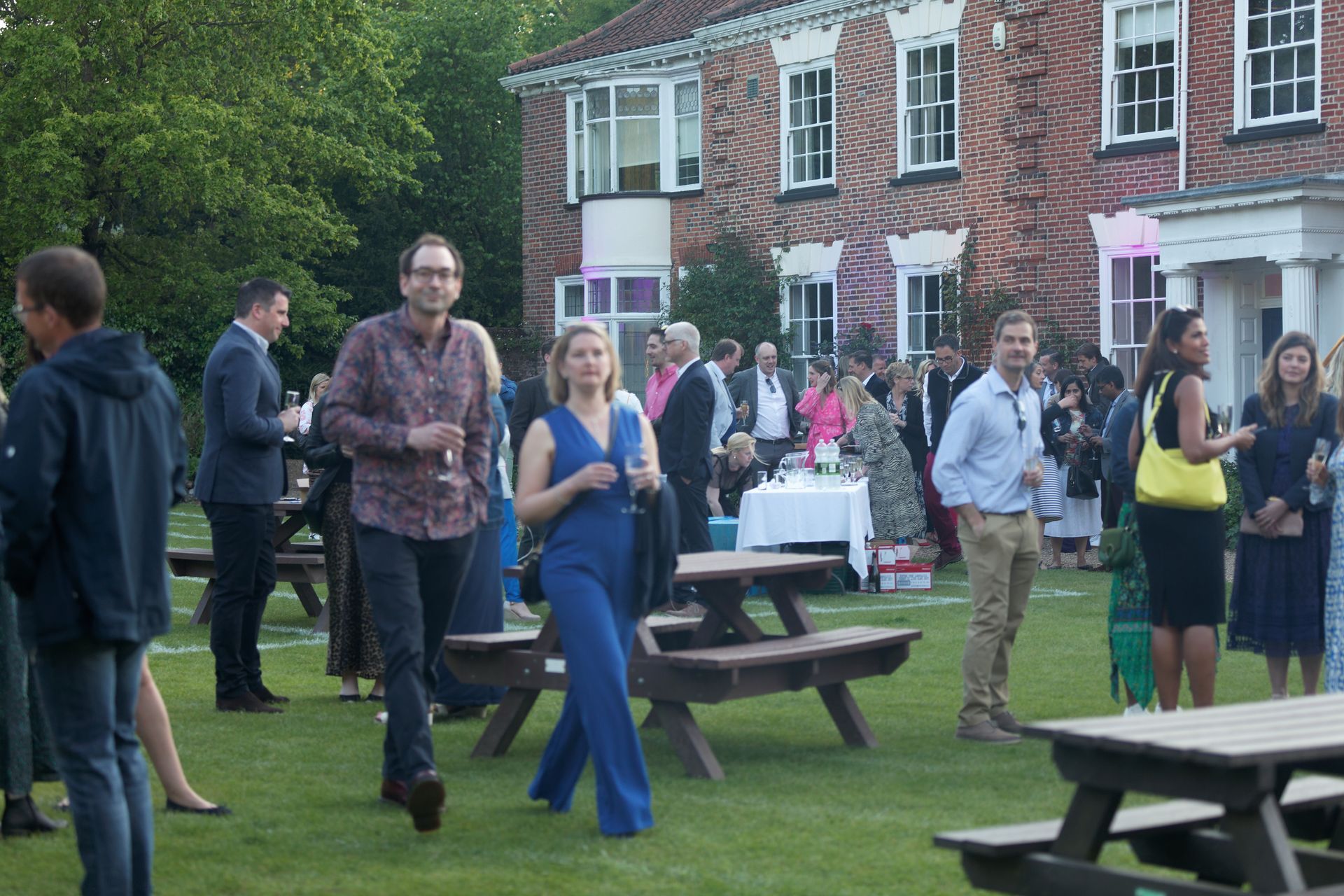 People at an outdoor event on grass, near a brick building. Some are socializing, others near picnic tables.