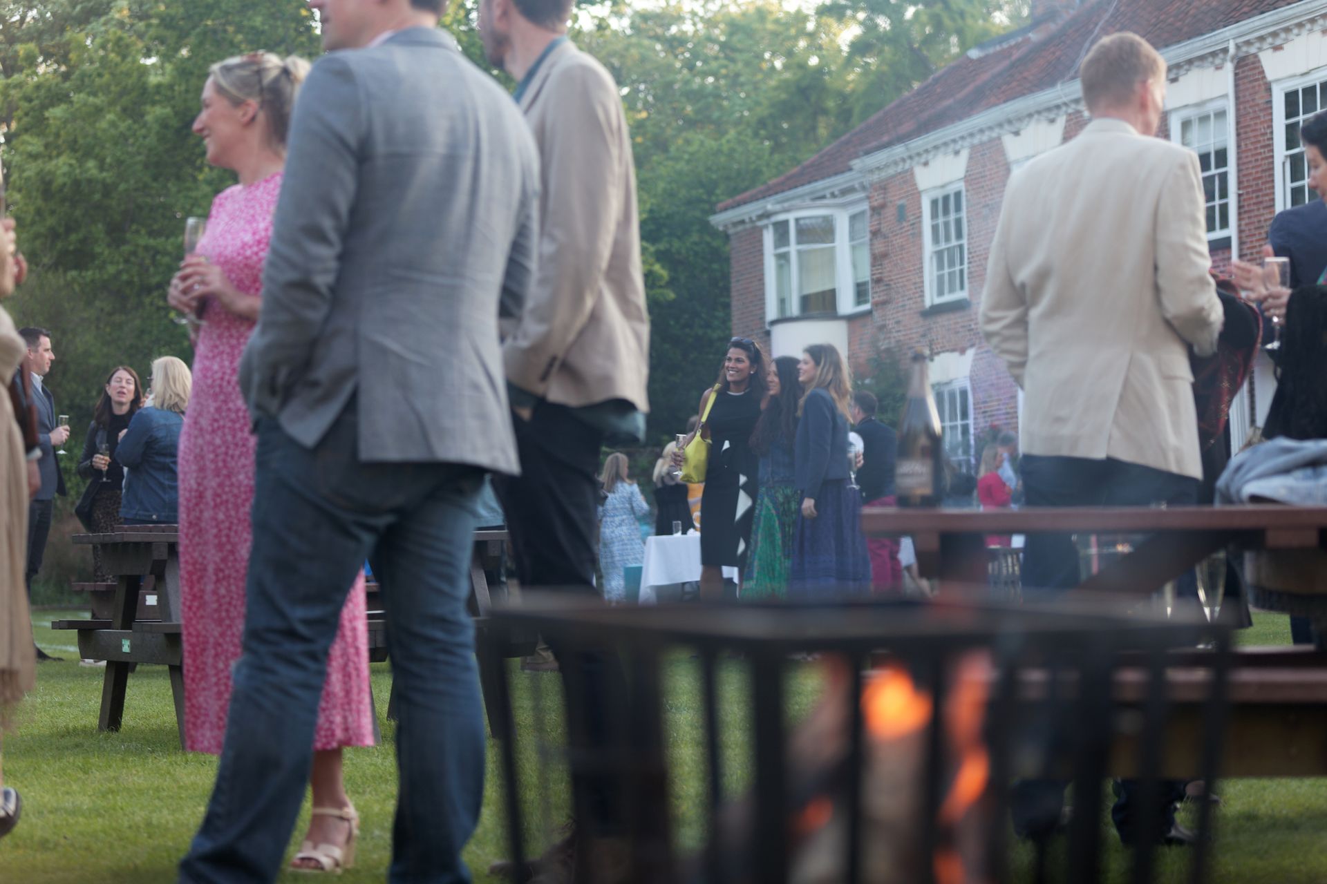 People at an outdoor gathering near a fire pit, house in the background, evening lighting.