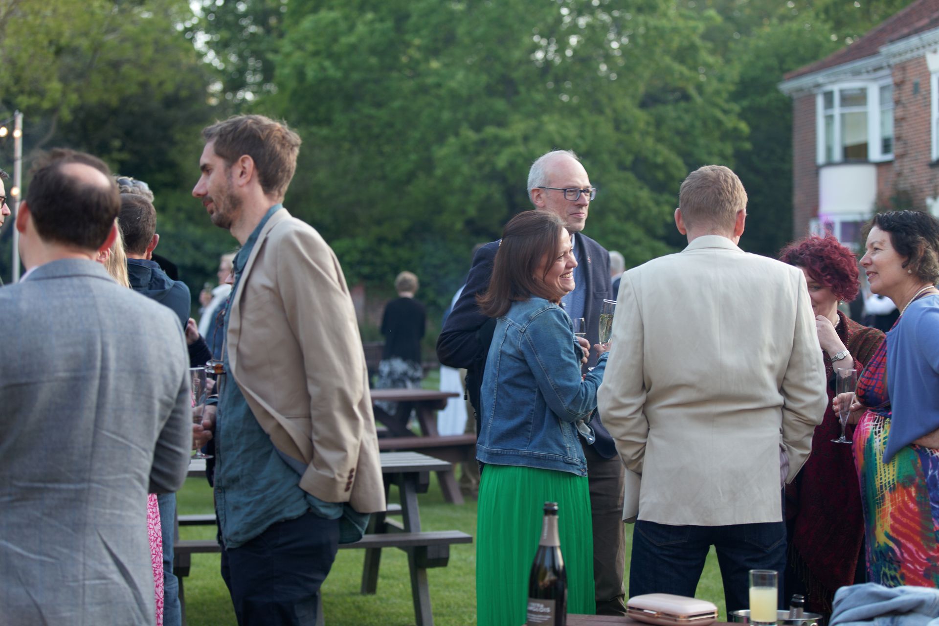 People socializing outdoors at an event on a lawn, some wearing suits and jackets, some smiling and talking.