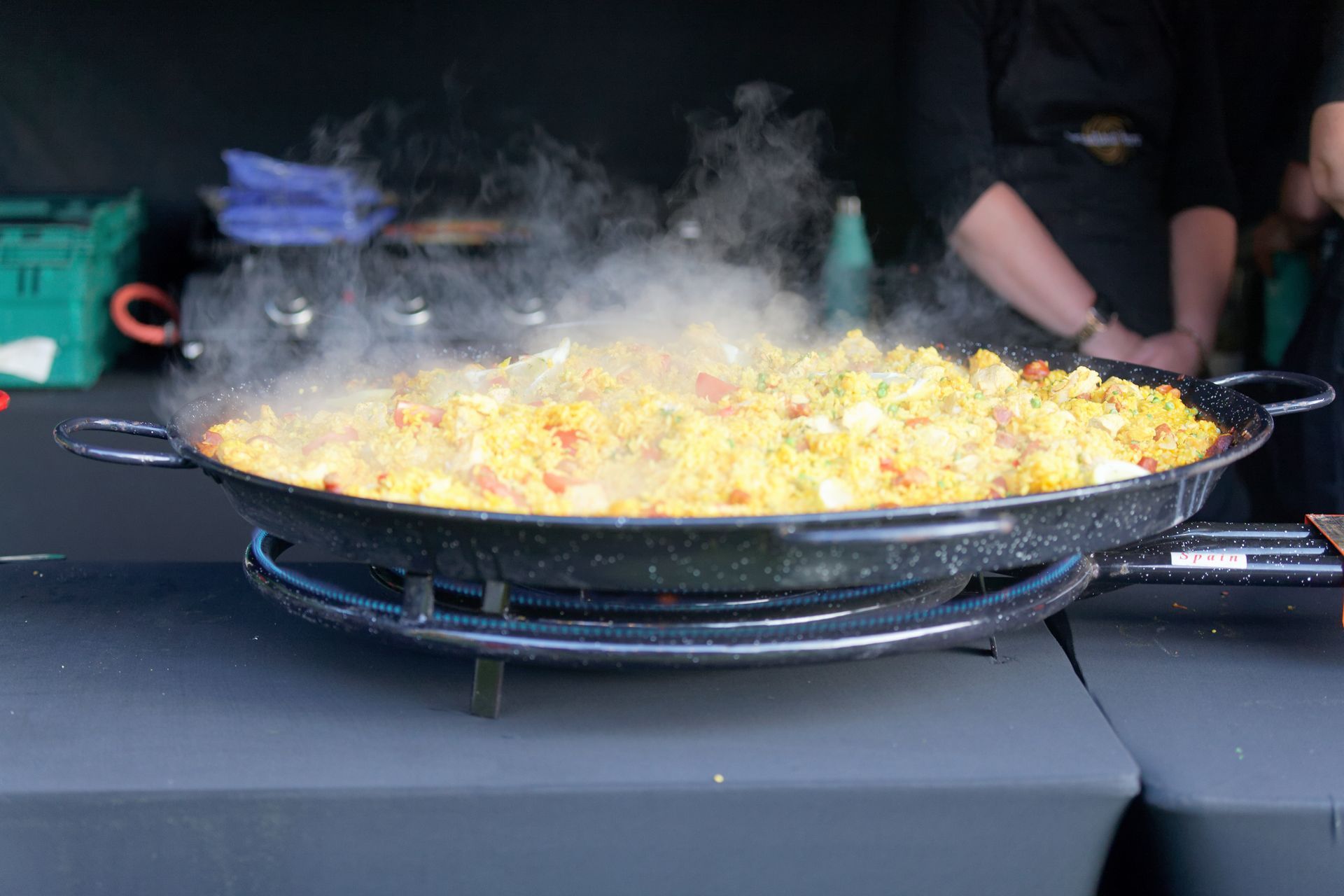 Paella cooking in a large, black pan, steaming over a gas burner; outdoor food vendor.