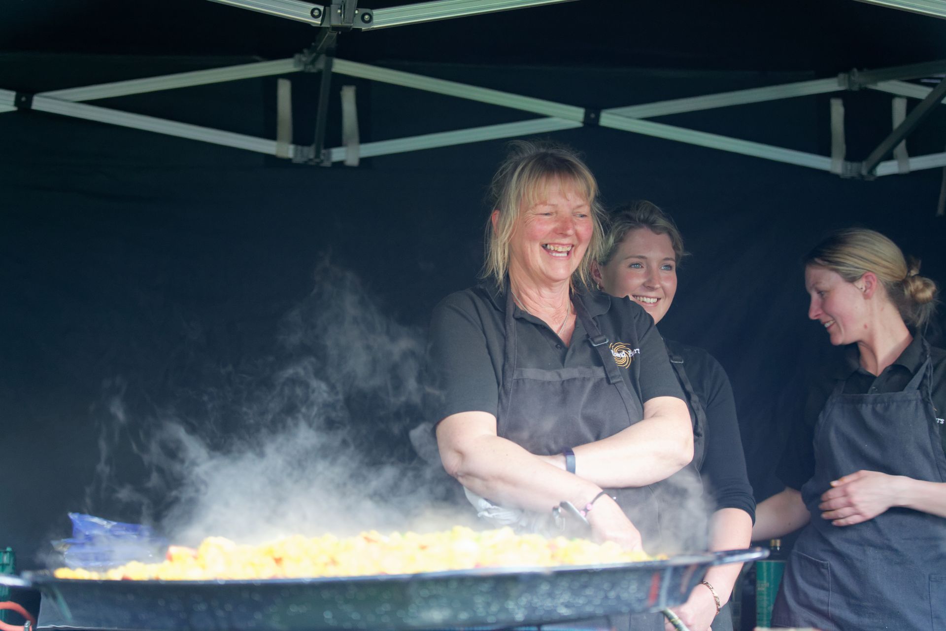 Three women at a food stall, smiling, with a large pan of steaming food. Black tent backdrop.