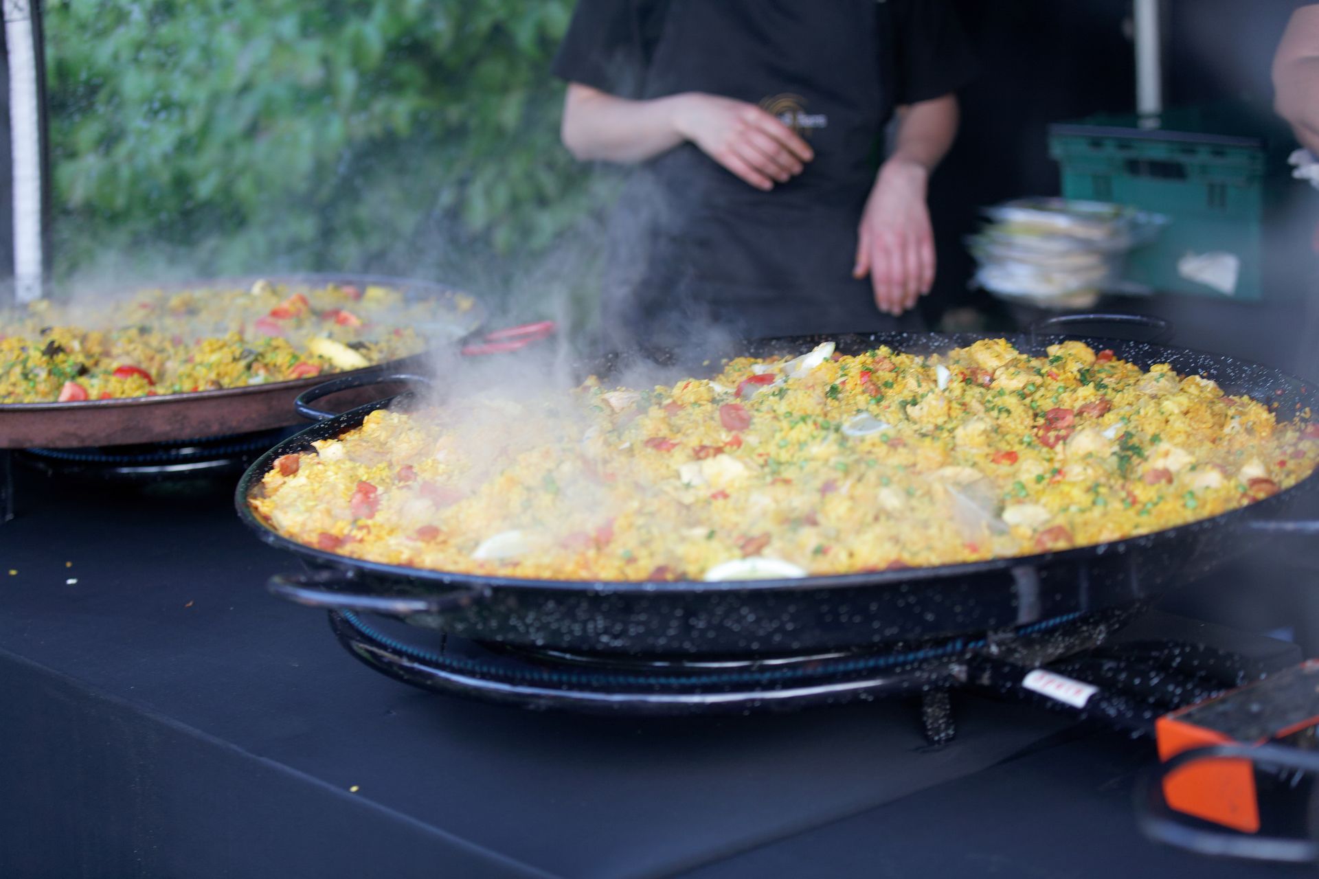 Two large pans of paella cooking, steam rising, at a food stall.