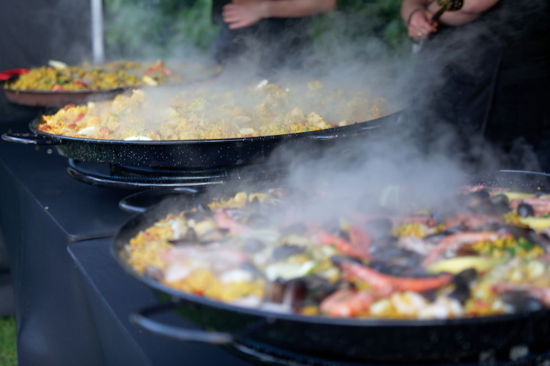 Paella cooking in large pans, steam rising. Seafood visible in a pan. Outdoor setting.