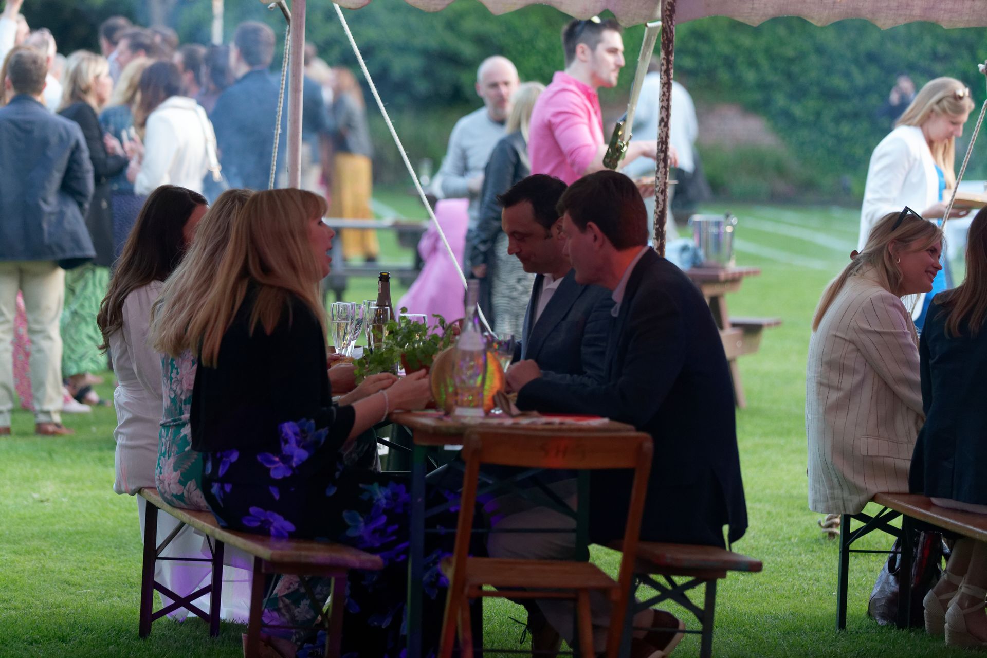 People gathered at outdoor event, seated at tables on a lawn. Some are eating and talking.