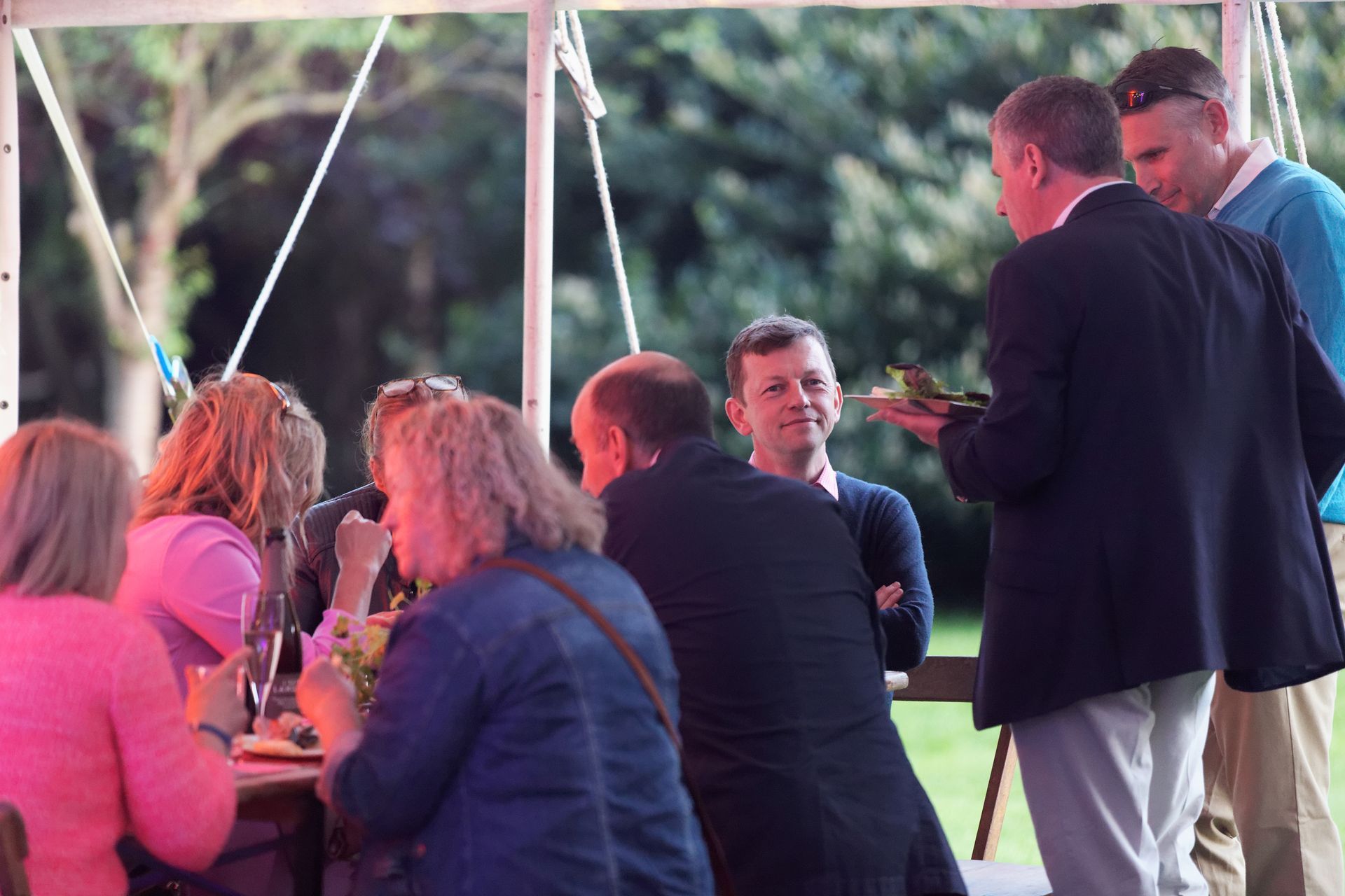 People at an outdoor event, under a tent. Some are eating, a man is serving food.