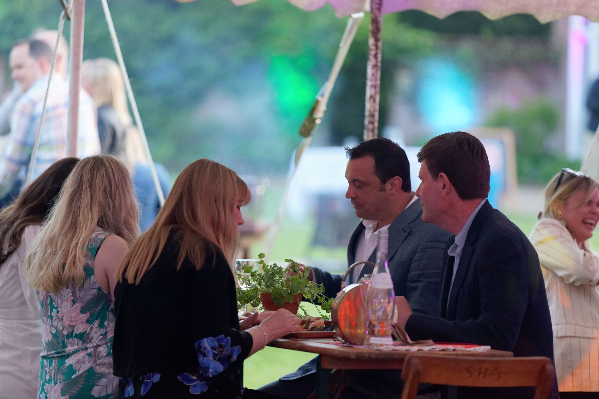 People seated at a table under a tent, conversing. A woman laughs in the background.