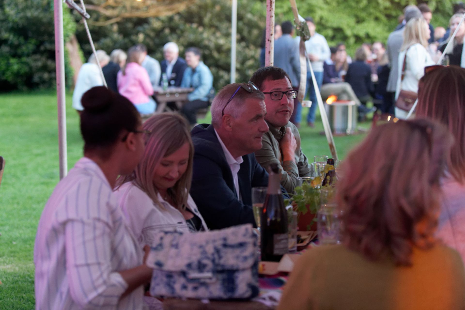 People seated at a table outdoors, socializing. Some are looking at others, smiling. Green grass and trees in the background.