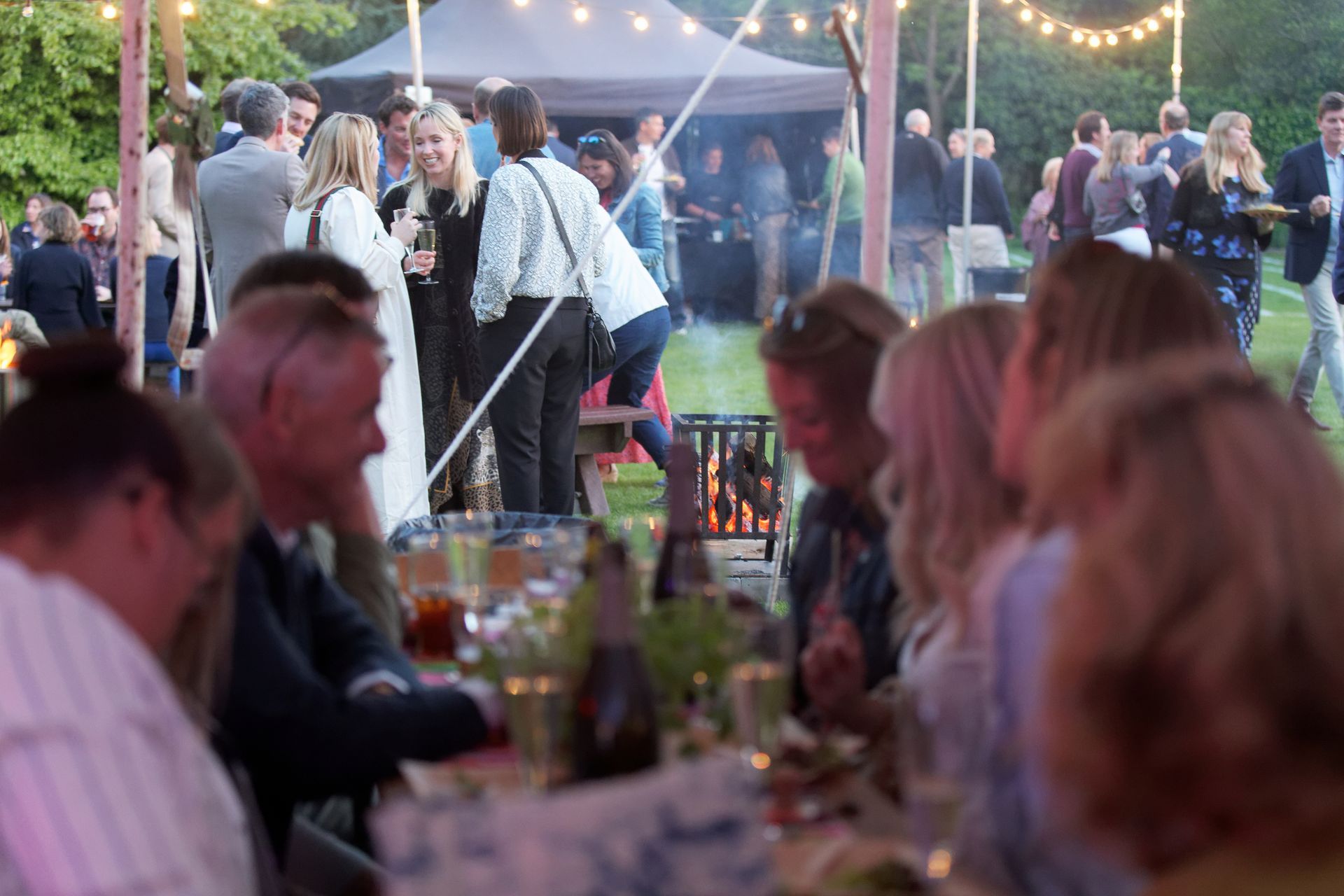 Guests at outdoor party, tables set, people mingling and eating, string lights, evening.