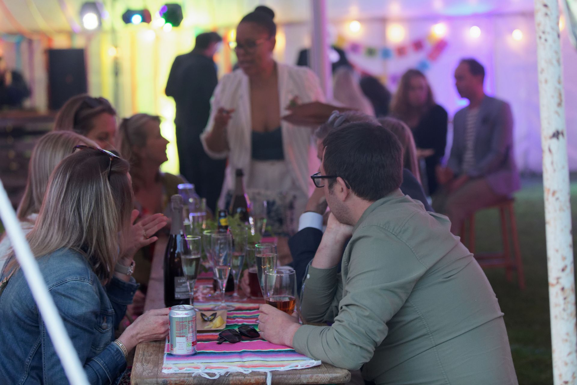 People at a party with drinks on a table, a person is speaking, colorful decorations, and string lights.