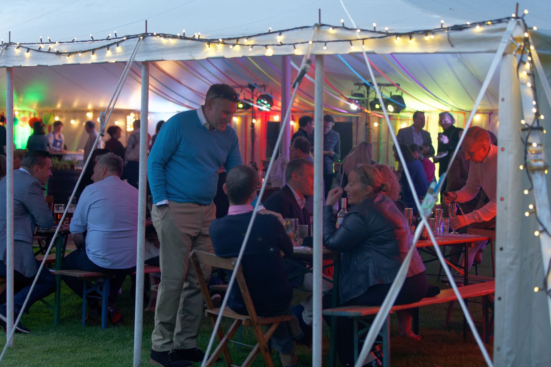 People socializing inside a tent lit with string lights. Man in blue sweater leans over a table.