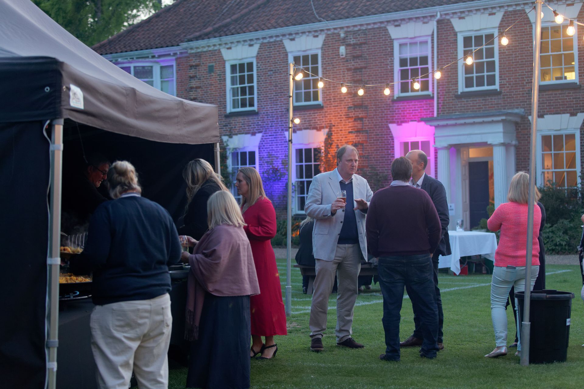 People at outdoor event in front of a brick building with food tent, string lights, and colorful up-lighting.