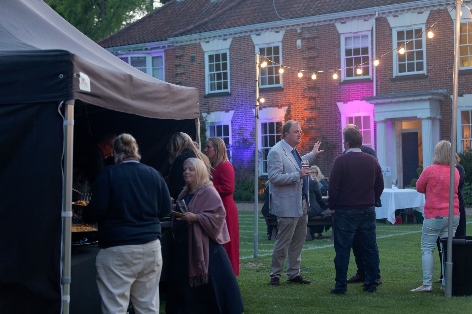 People at outdoor event near a brick building, with string lights, a tent, and colorful lighting.