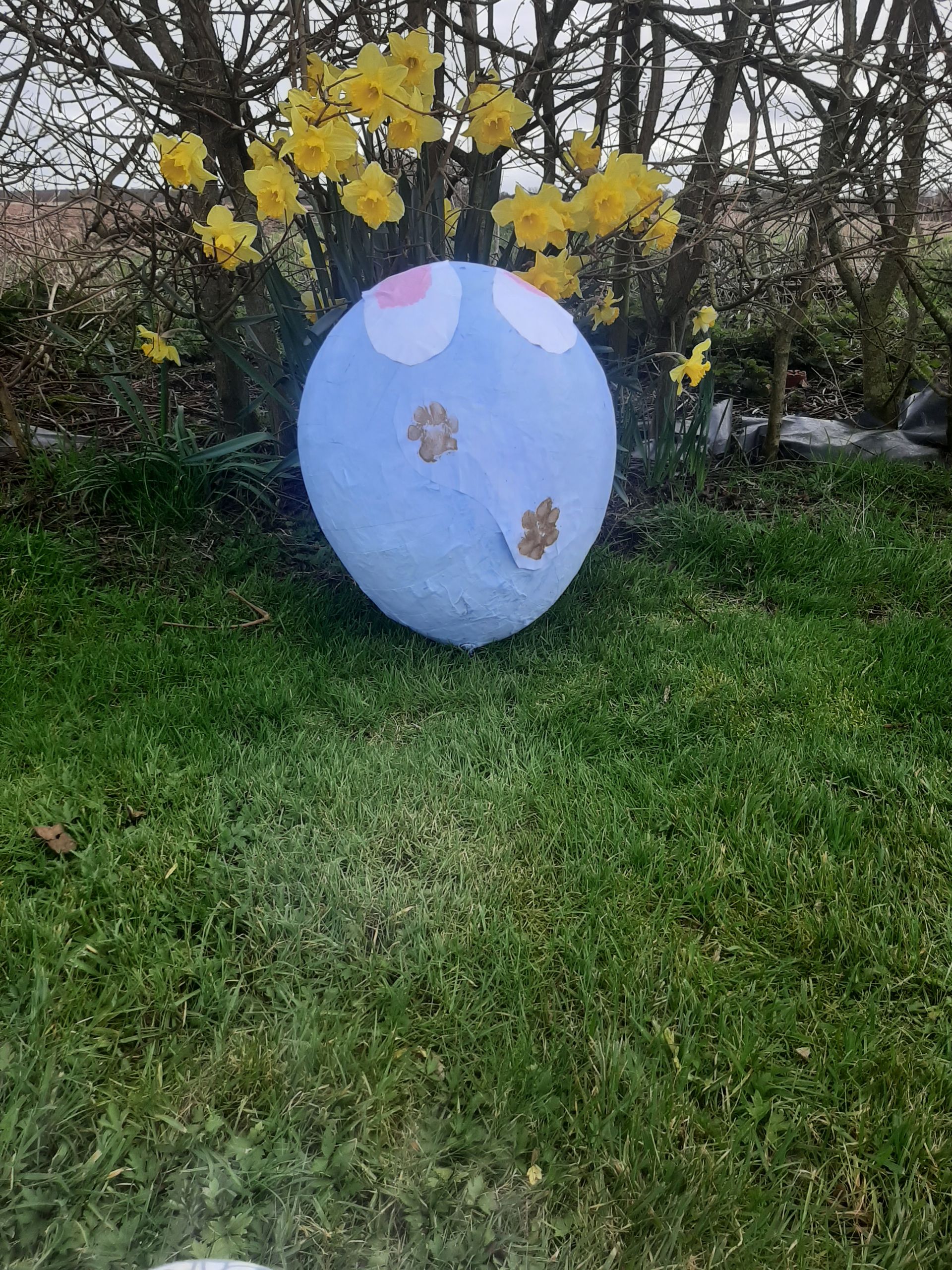 Large, light blue Easter egg decorated with pink and brown spots, sits on green grass in front of yellow flowers.
