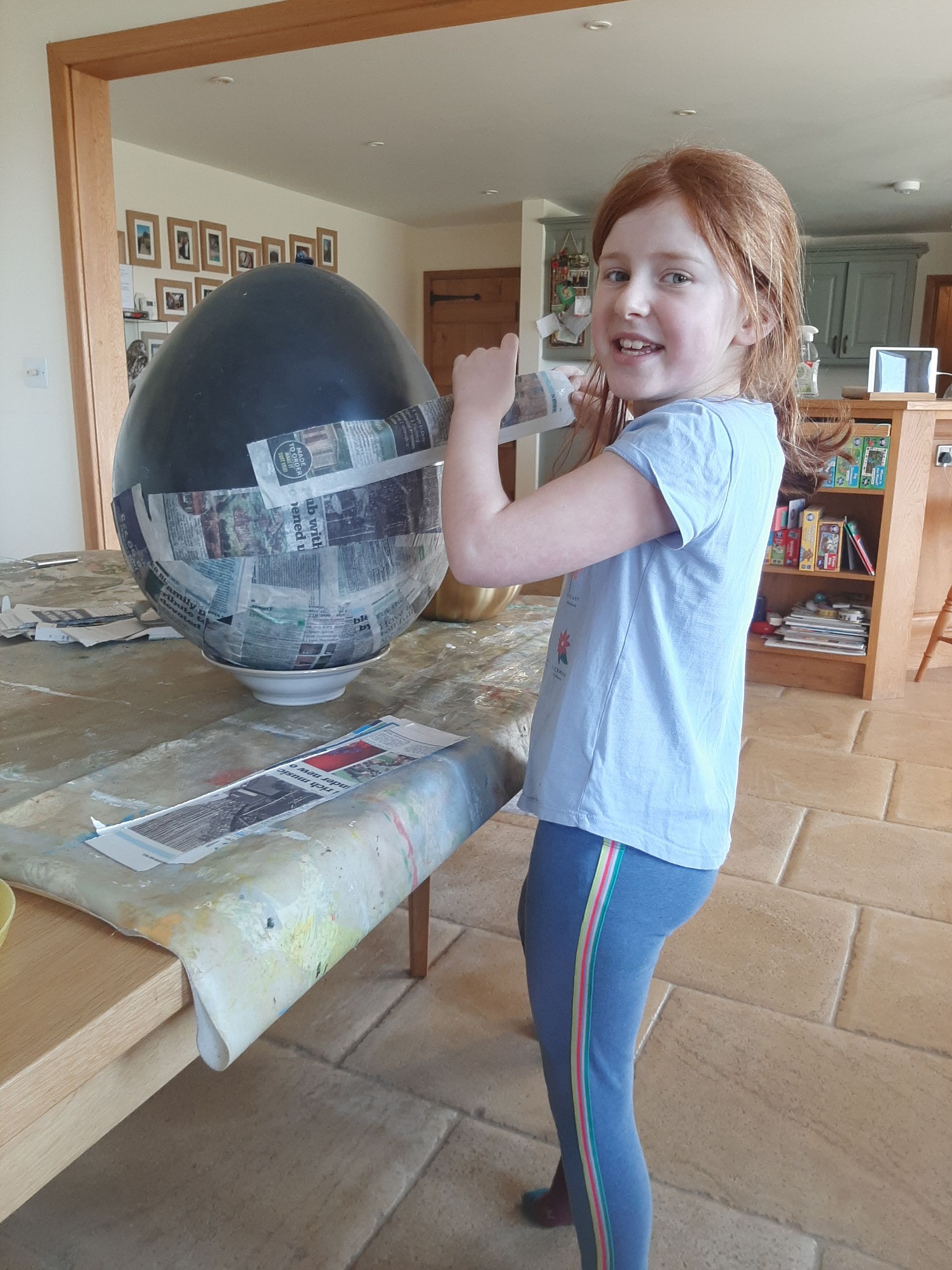 Young girl smiles next to a large, paper-mâché egg. Indoors, with newspaper scraps on a table.