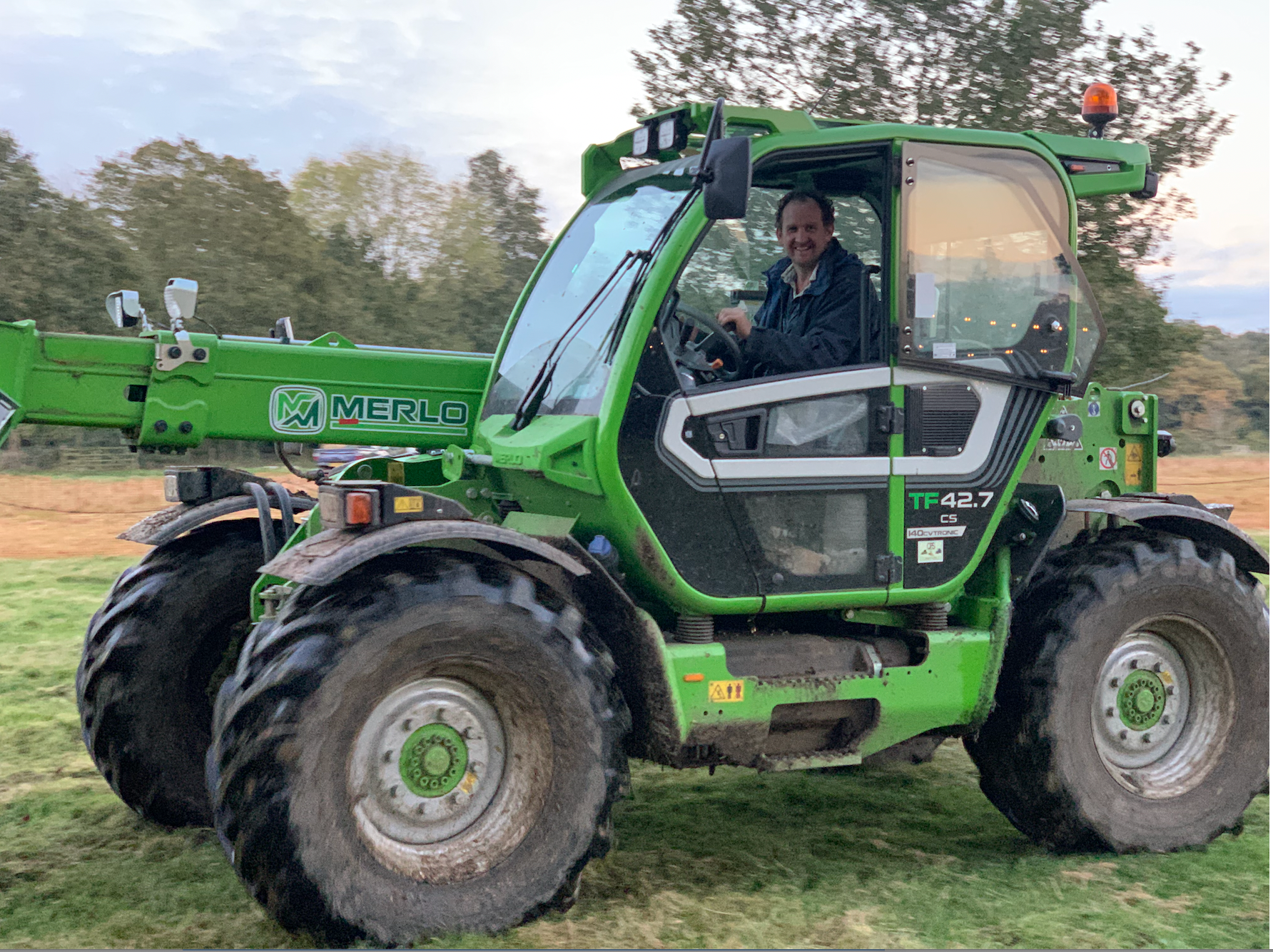 Man driving a green telescopic handler in a field, smiling at the camera.