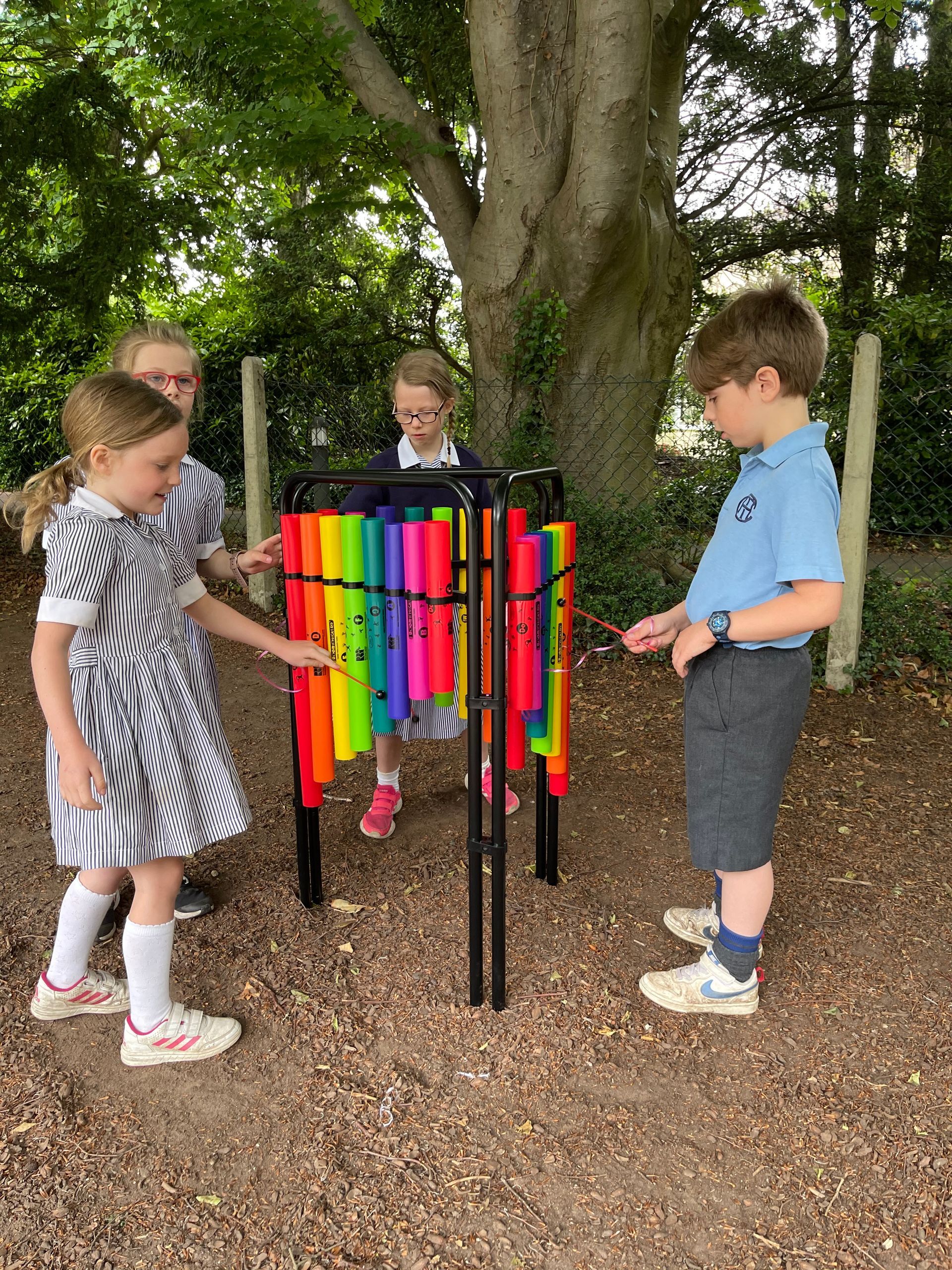 Children playing a rainbow-colored outdoor xylophone, in a park-like setting with trees.
