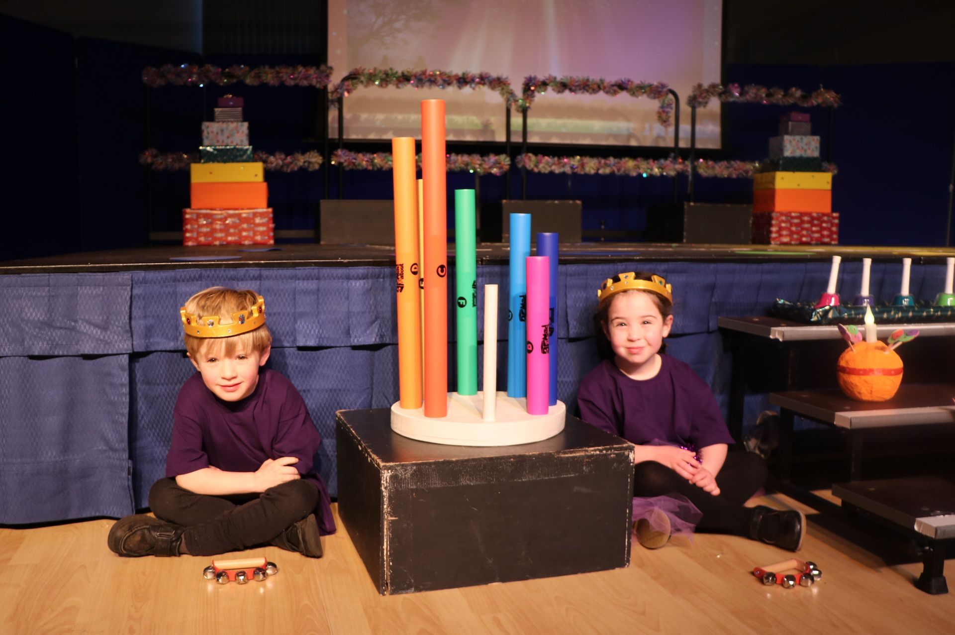 Two children in crowns and purple shirts sit on stage with a colorful musical instrument.