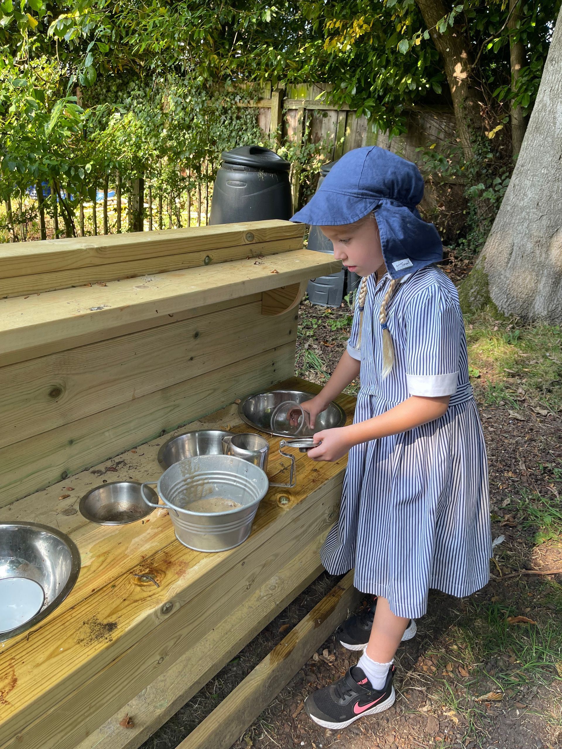 Girl in a hat and dress playing with bowls and sand on a wooden play structure outdoors.