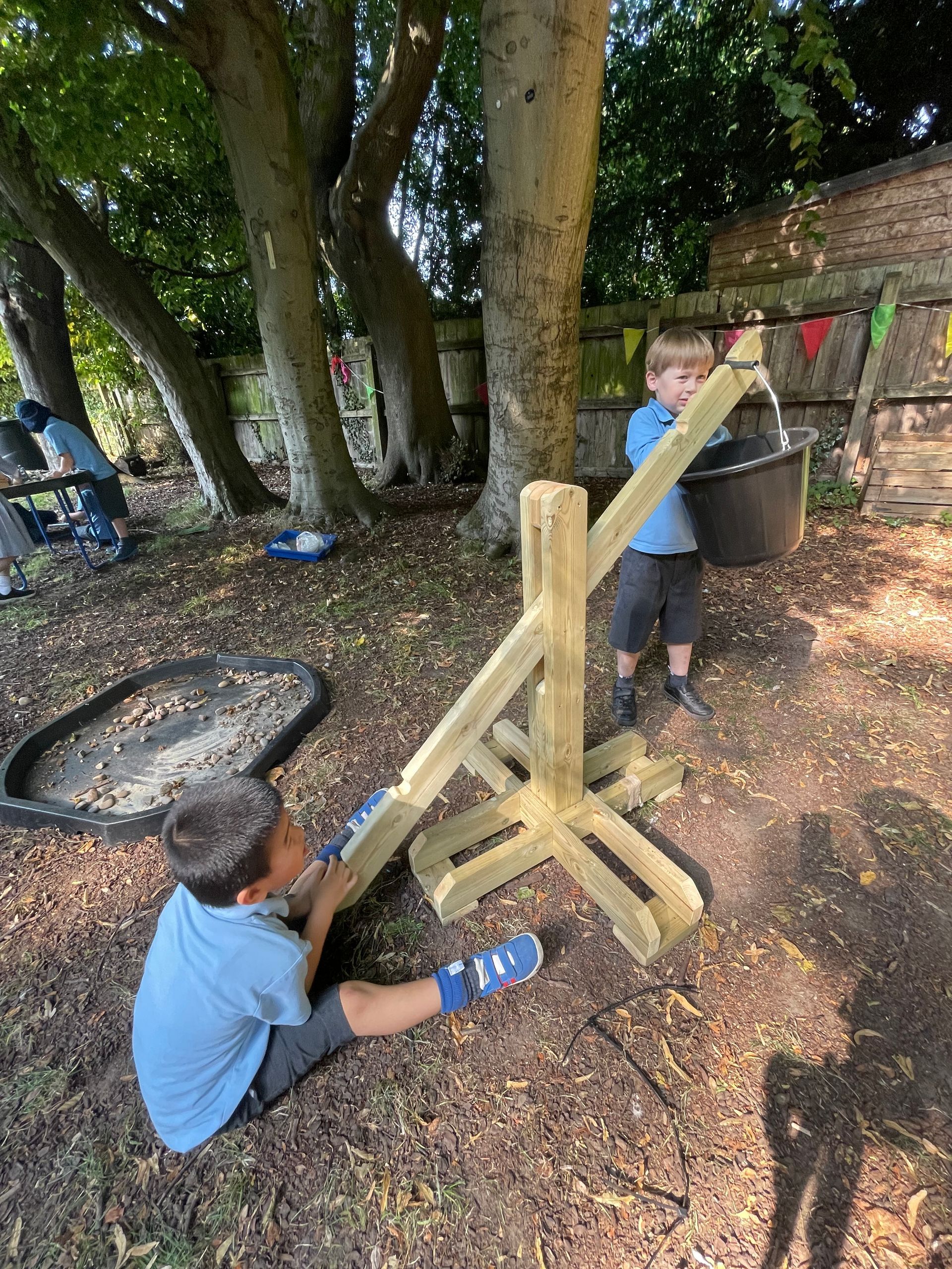 Two children in a yard using a wooden catapult, one loads a bucket, another pulls the lever.