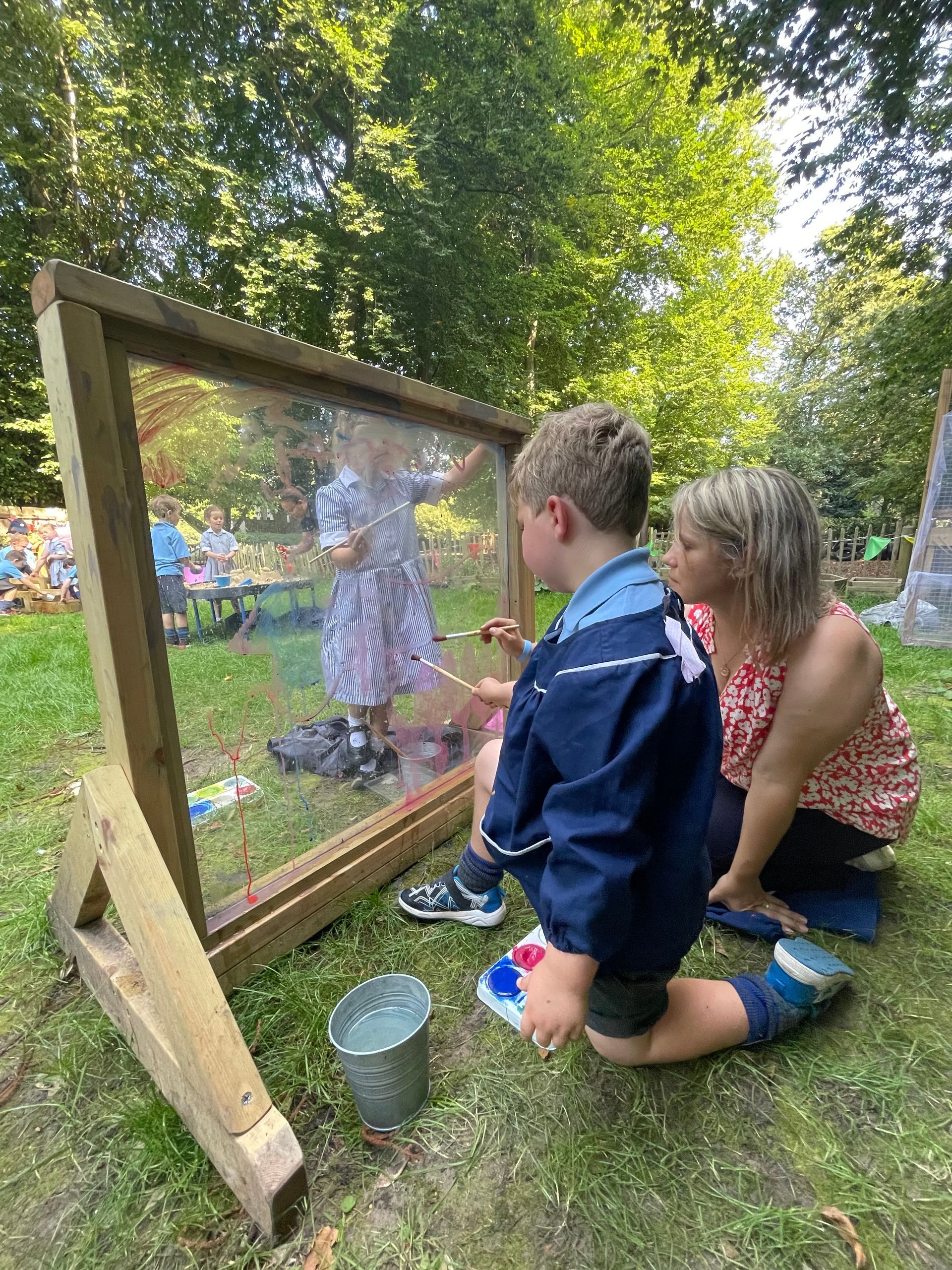 A young boy and adult painting on a clear board outdoors. Another child visible in background.
