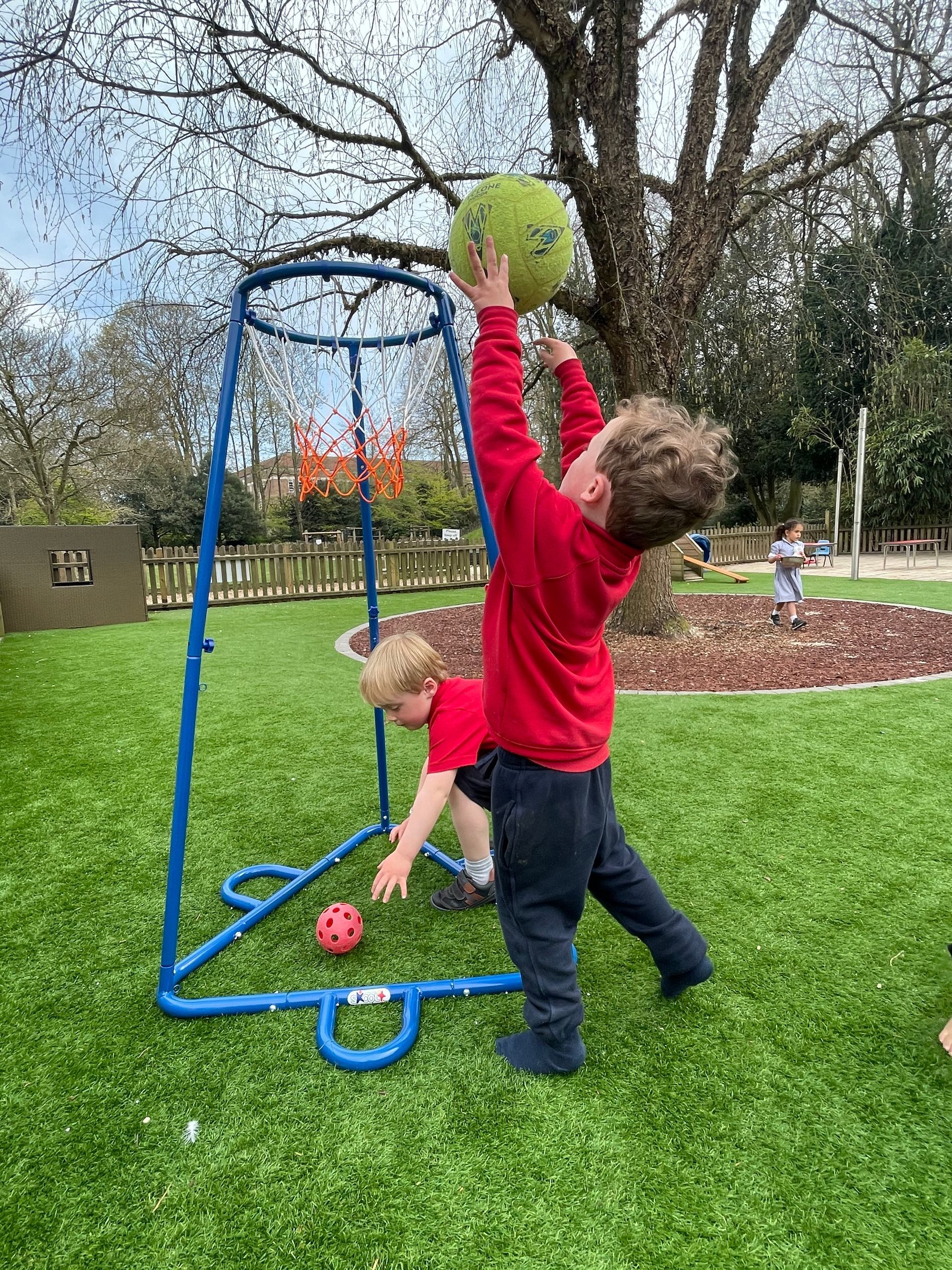 Two young children playing a ball game on green turf near a blue hoop structure, one with arms raised.