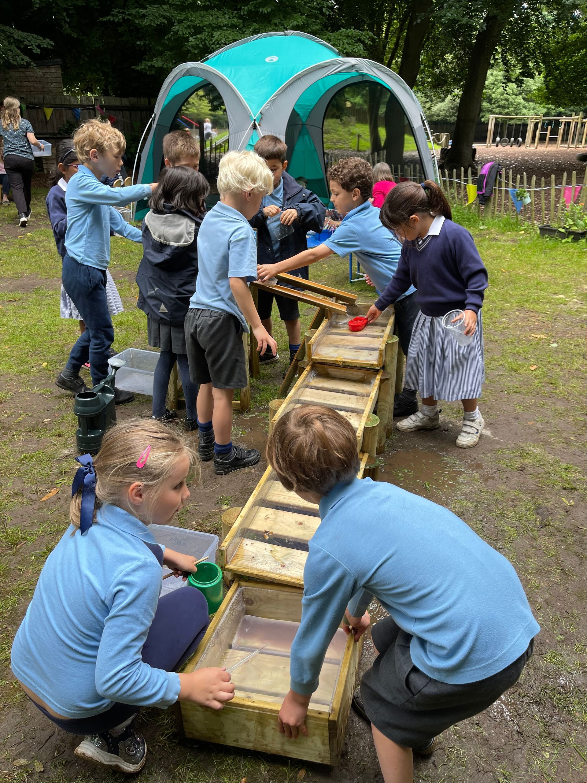 Children playing with water in wooden troughs, outdoors. Light blue shirts, school uniforms.