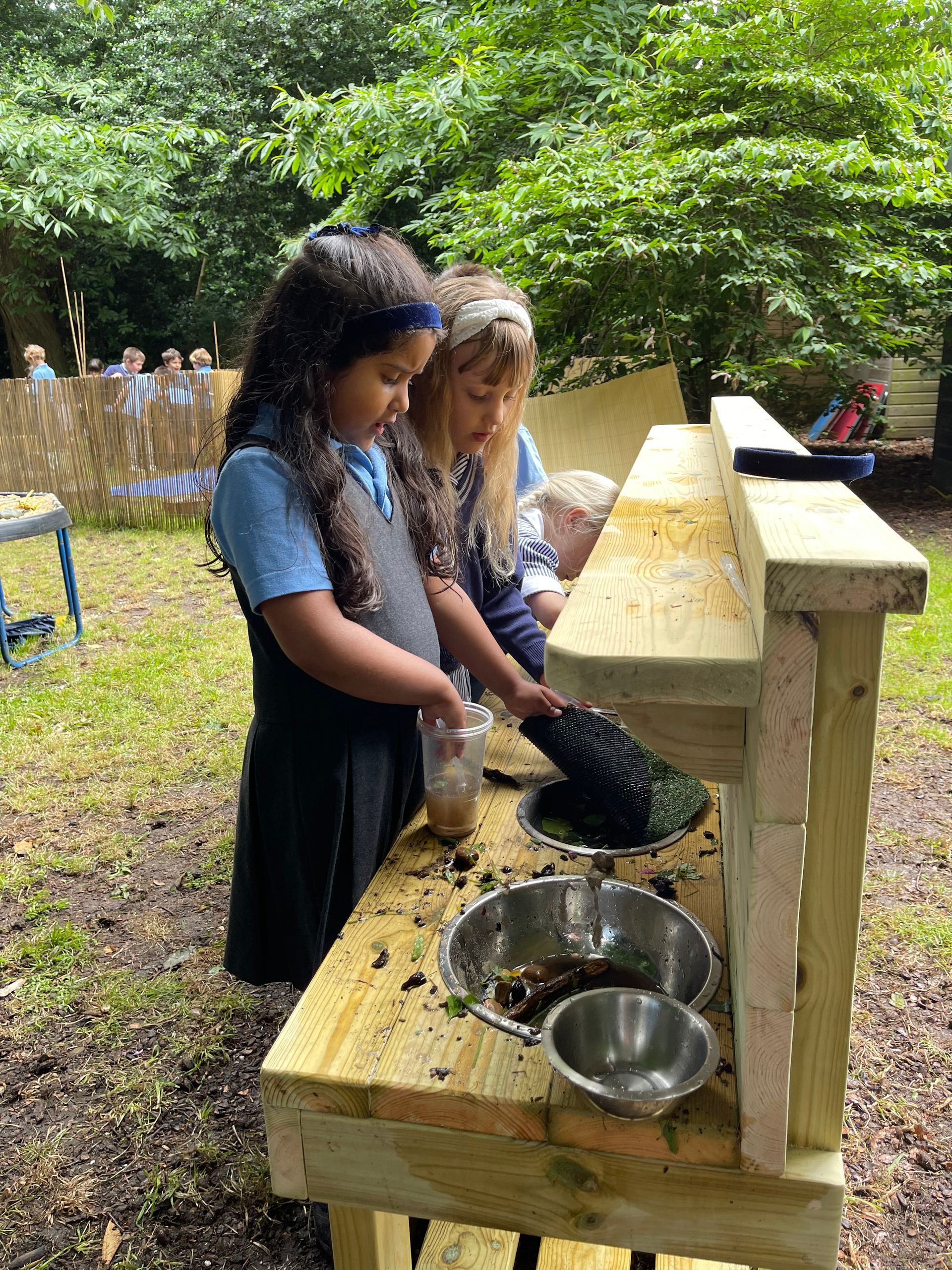 Two girls in school uniforms use a mud kitchen outdoors; one strains, the other watches.