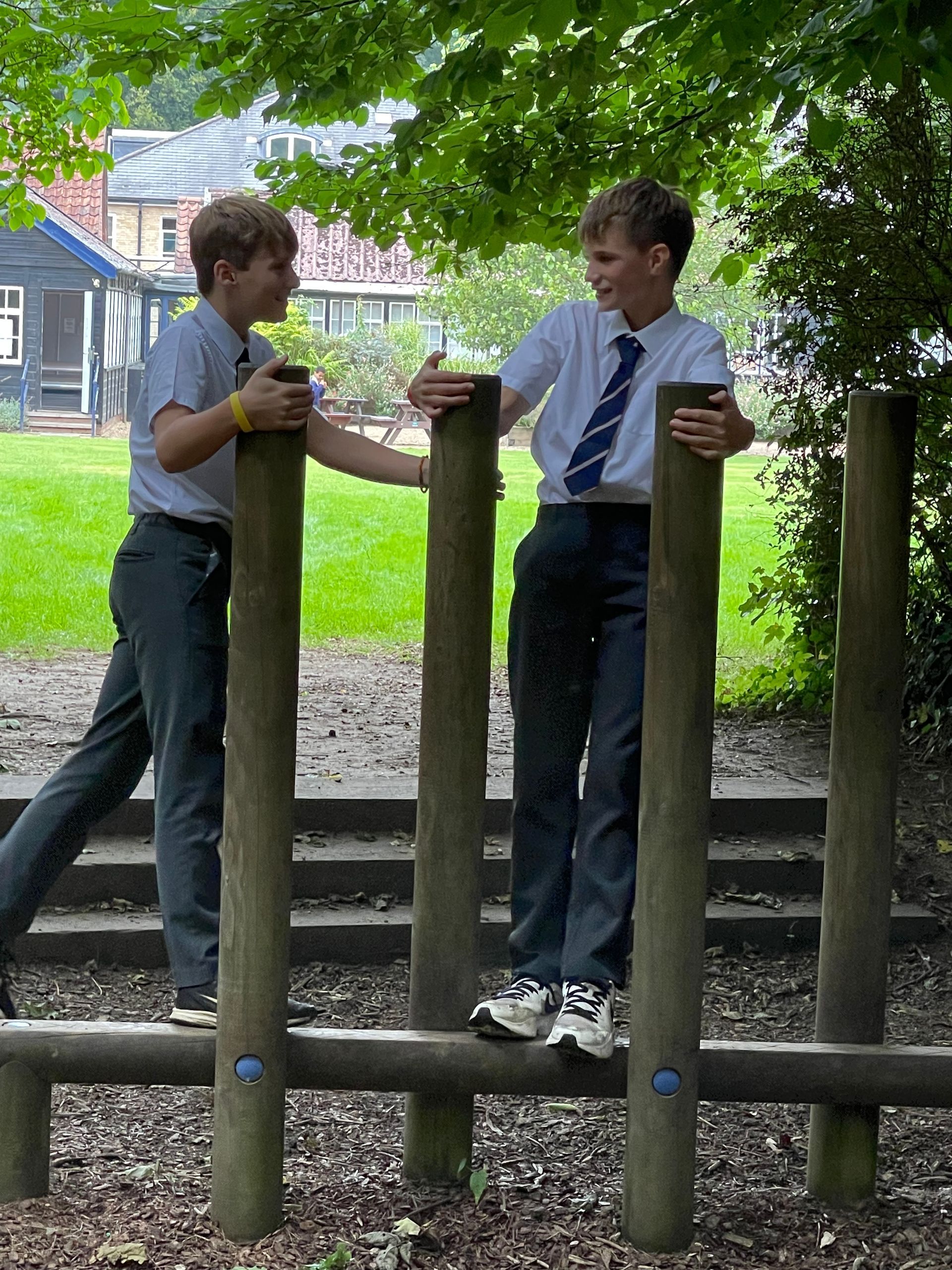 Two boys in school uniforms balance on wooden posts in a grassy outdoor area.