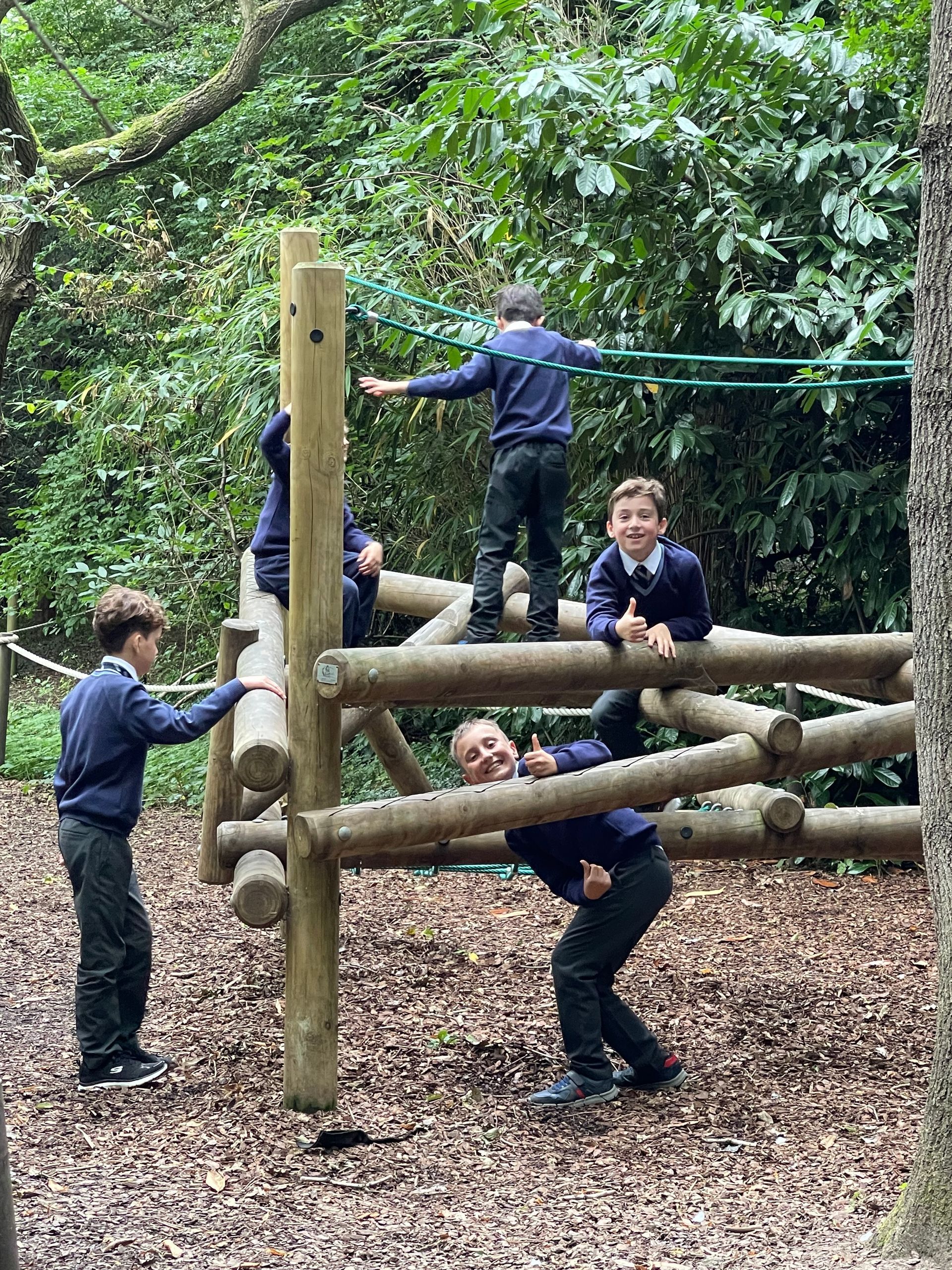 Boys in school uniforms play on a wooden climbing structure in a wooded area.