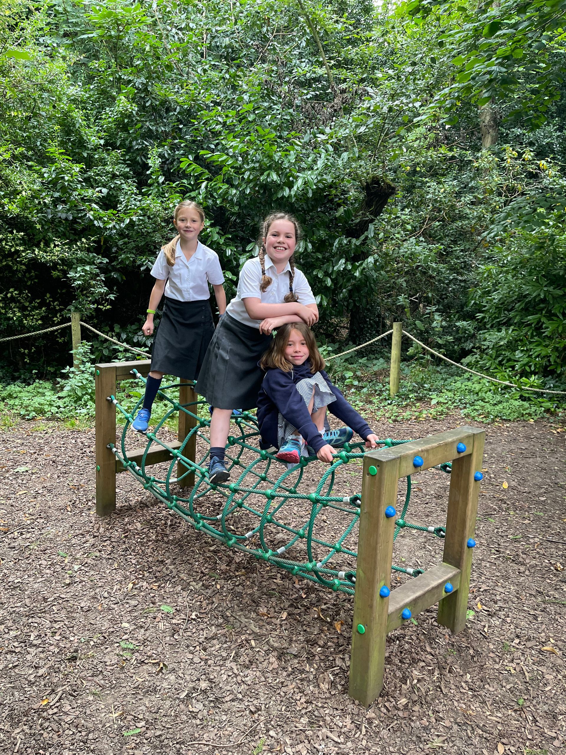 Three children on a rope bridge in a wooded playground. One is smiling, others looking at camera.